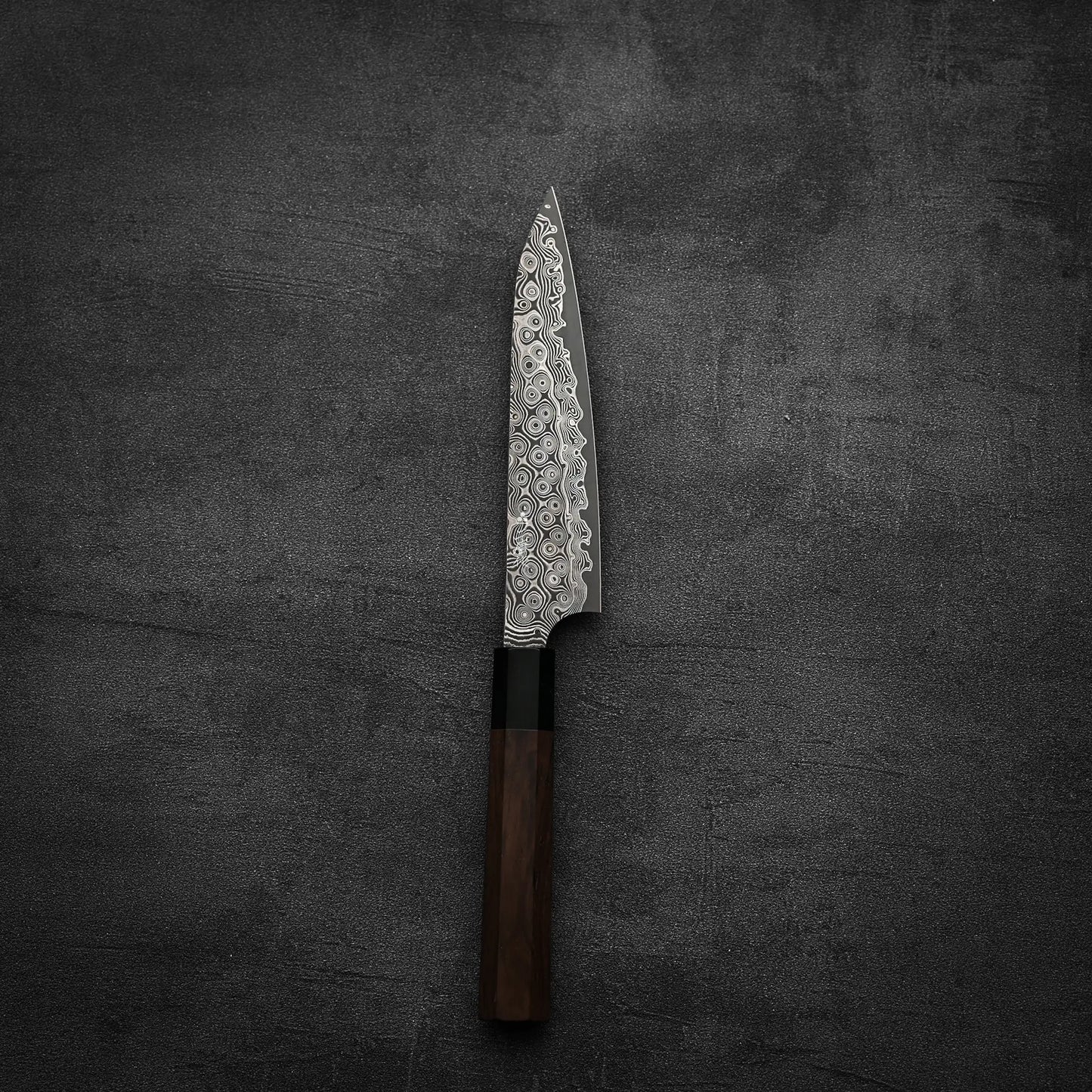 The Nigara Anmon SG2 damascus petty knife 150mm by Nigara Hamono, featuring a patterned blade and dark wooden handle, is shown vertically with the blade pointing up on a dark textured surface.