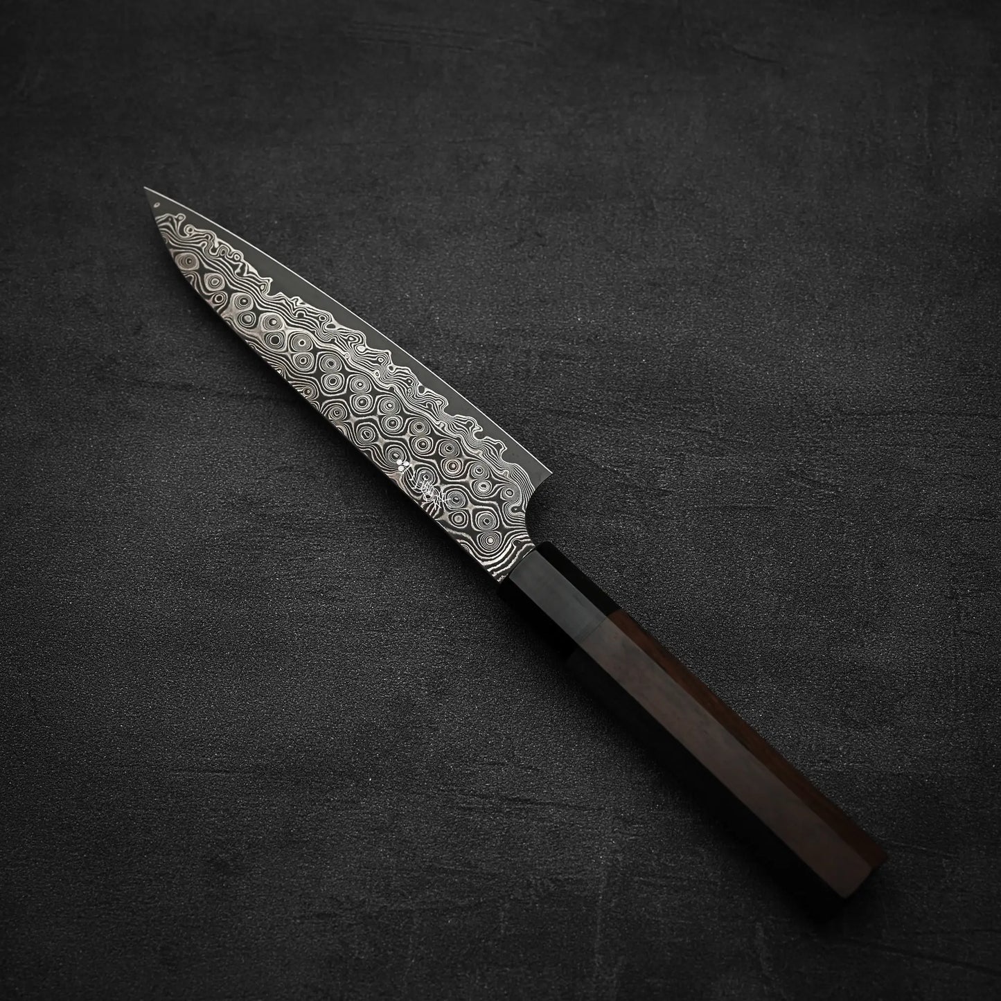 The Nigara Hamono Nigara Anmon SG2 Damascus petty knife (150mm), featuring a patterned blade and dark wood handle, rests on a textured dark gray surface.