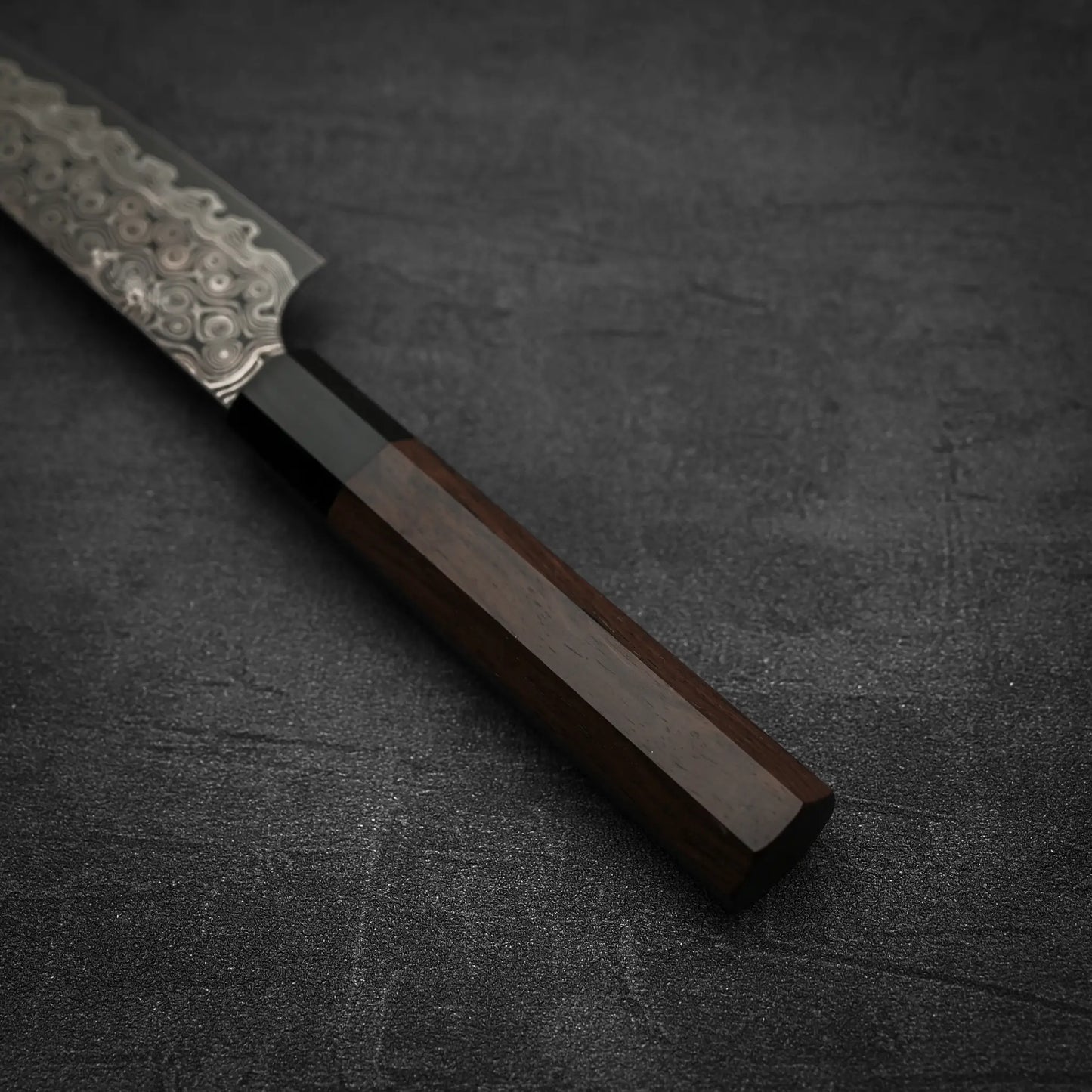 A close-up of the Nigara Hamono Nigara Anmon SG2 damascus petty knife 150mm, featuring a patterned steel blade and dark wooden handle, displayed on a dark textured surface.
