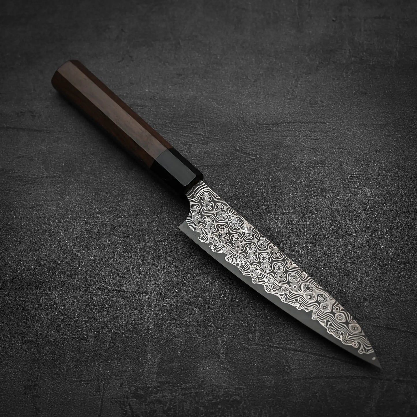 The Nigara Hamono Nigara Anmon SG2 damascus petty knife 150mm, featuring a dark wooden handle and patterned blade, rests on a textured dark surface.