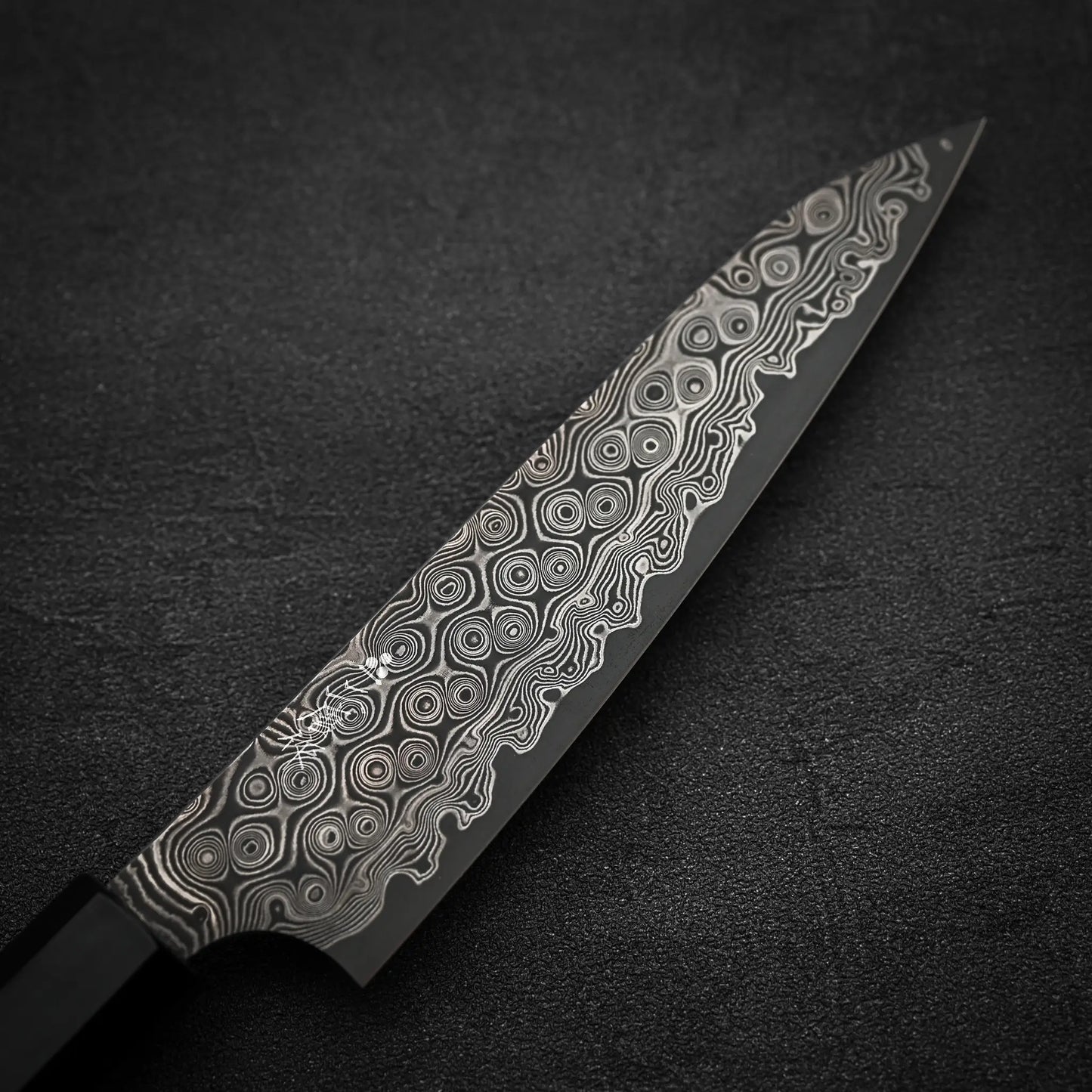 A close-up of the Nigara Hamono Nigara Anmon SG2 Damascus petty knife 150mm, showcasing its intricate swirling patterned steel blade on a dark textured surface.