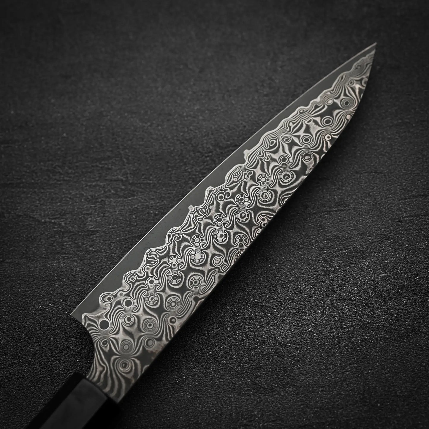 A close-up of the Nigara Anmon SG2 damascus petty knife 150mm by Nigara Hamono, featuring a handcrafted patterned Damascus steel blade with intricate swirling designs, placed on a dark textured surface.
