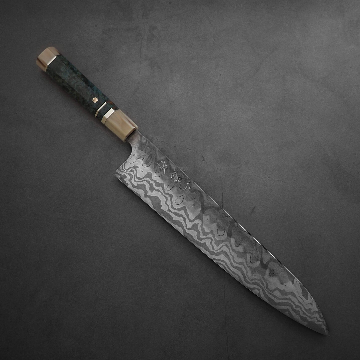 The Nigara Hamono Nigara aogami#2 + shirogami#2 coreless honyaki gyuto 300mm, with a patterned Damascus blade and dark handle with metallic accents, rests on a dark, textured surface.