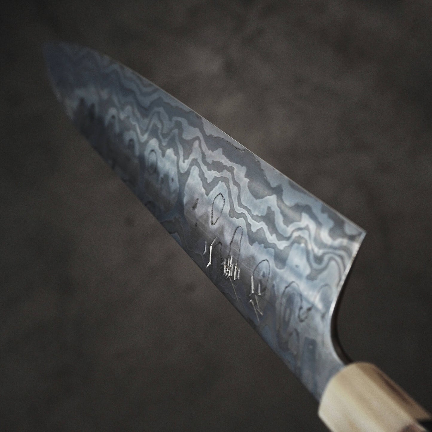 A close-up of the Nigara Hamono Nigara aogami#2 + shirogami#2 coreless honyaki gyuto 300mm, showing its Damascus-patterned blade, etched characters by the bolster, and wooden handle against a dark, blurred background.