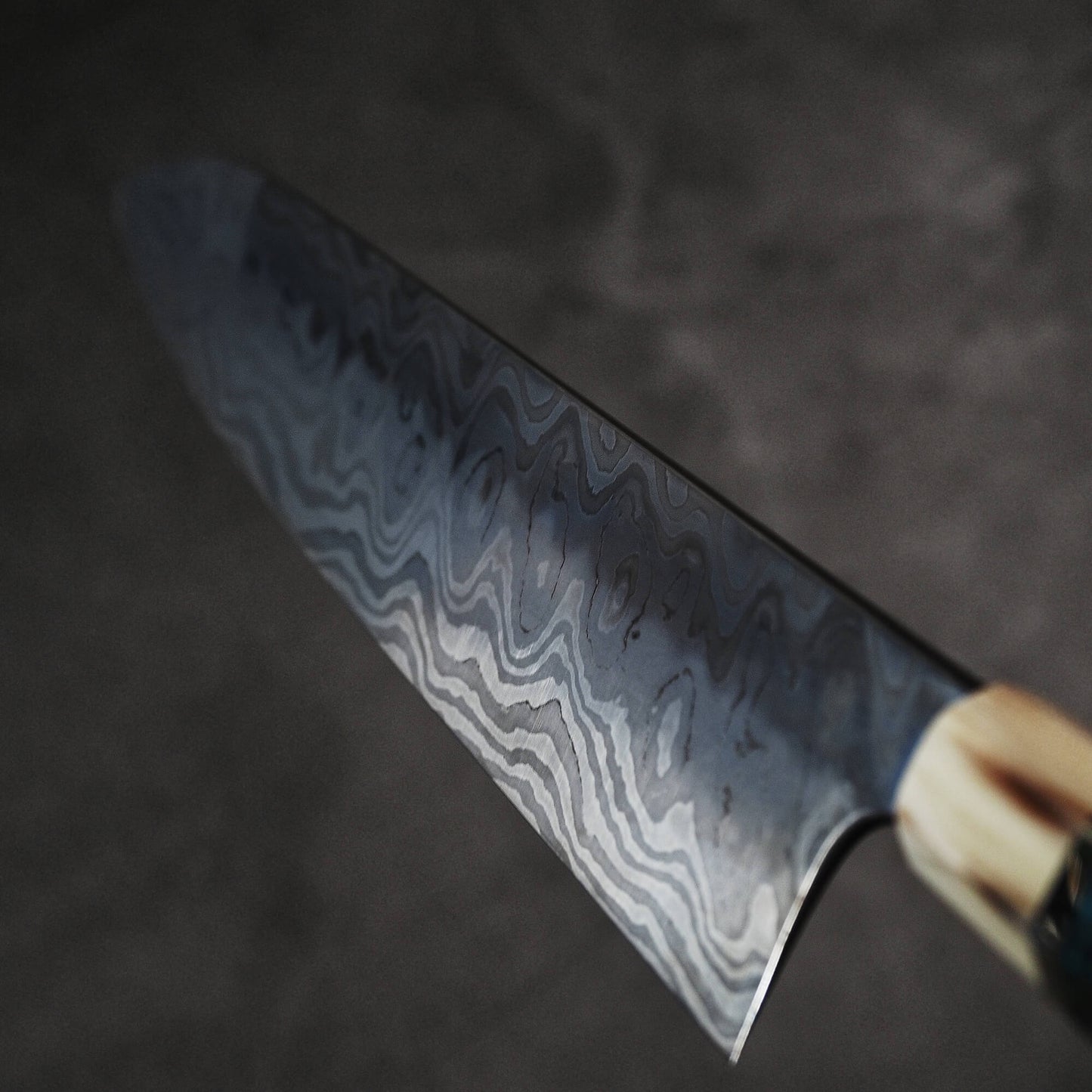A close-up of the Nigara Hamono Nigara aogami#2 + shirogami#2 coreless honyaki gyuto 300mm showcases its intricate wavy blade pattern, echoing Japanese swordsmith artistry, against a dark, blurred backdrop.