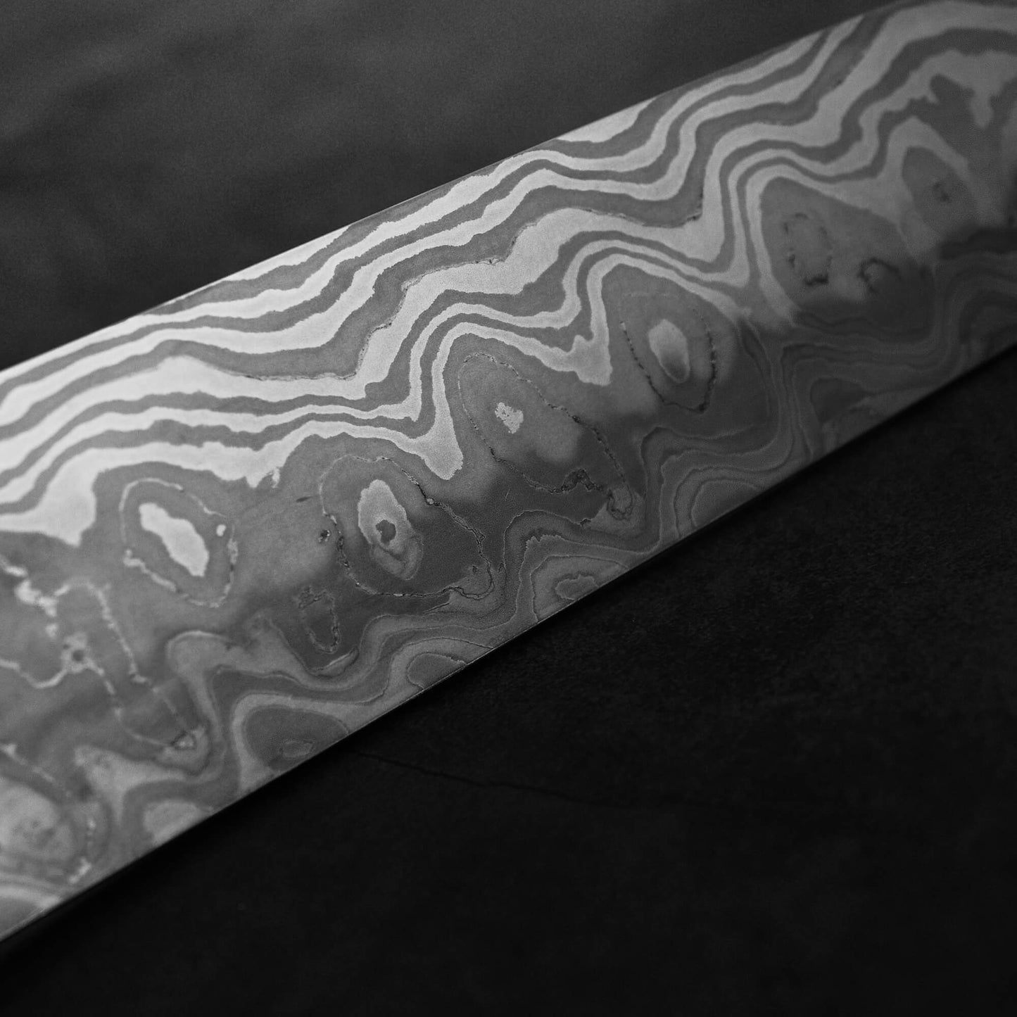 Black-and-white close-up of the Nigara Hamono Nigara aogami#2 + shirogami#2 coreless honyaki gyuto 300mm shows its intricate wavy patterns, inspired by Japanese swordsmithing, against a dark, textured background.
