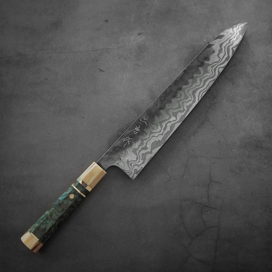 A Nigara Hamono Nigara aogami#2 + shirogami#2 coreless honyaki gyuto 300mm with a patterned blade and wooden handle featuring green and gold accents rests on a dark gray textured surface.