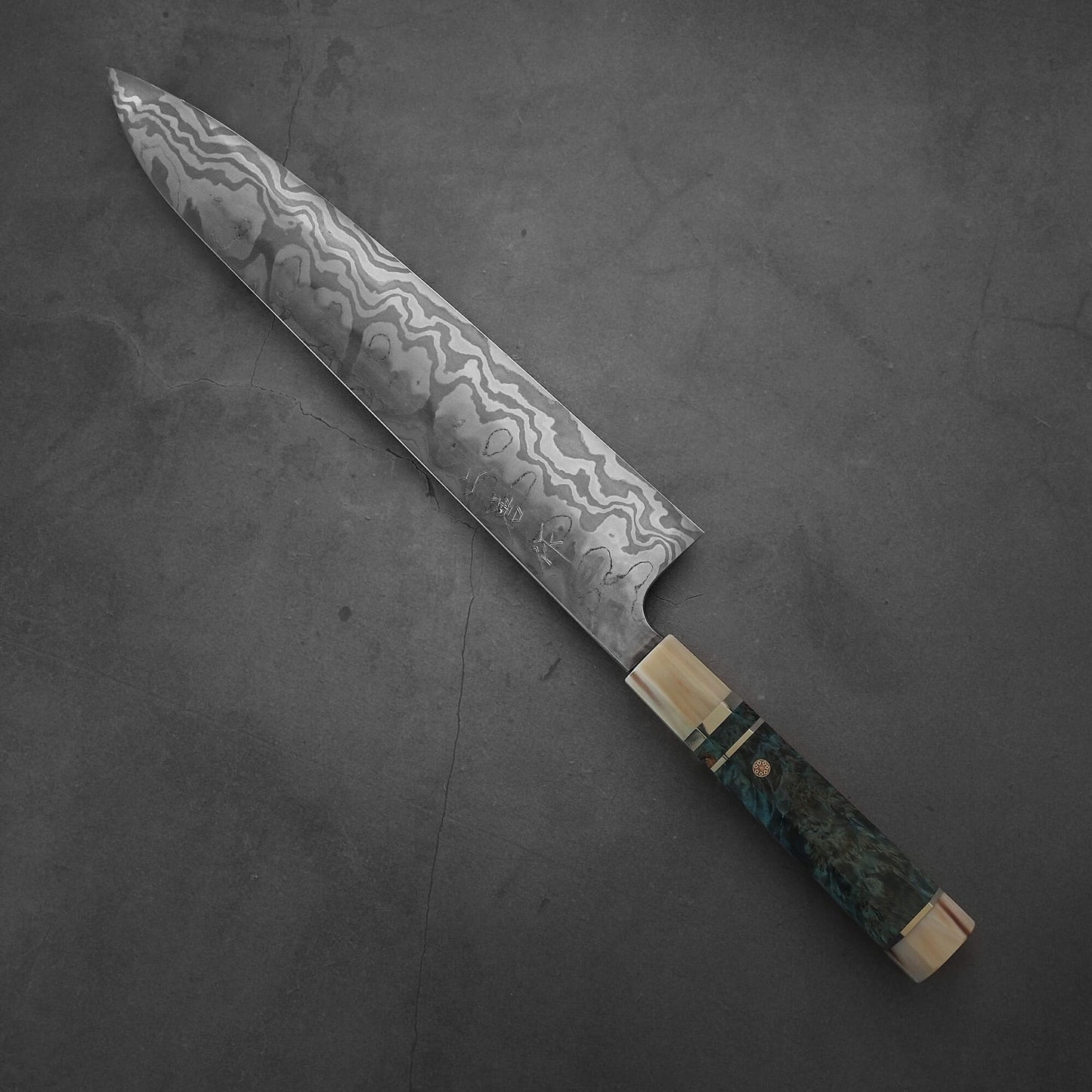 The Nigara Hamono Nigara aogami#2 + shirogami#2 coreless honyaki gyuto 300mm features a patterned blade and a dark wood and brass handle, showcased on a dark gray textured surface.