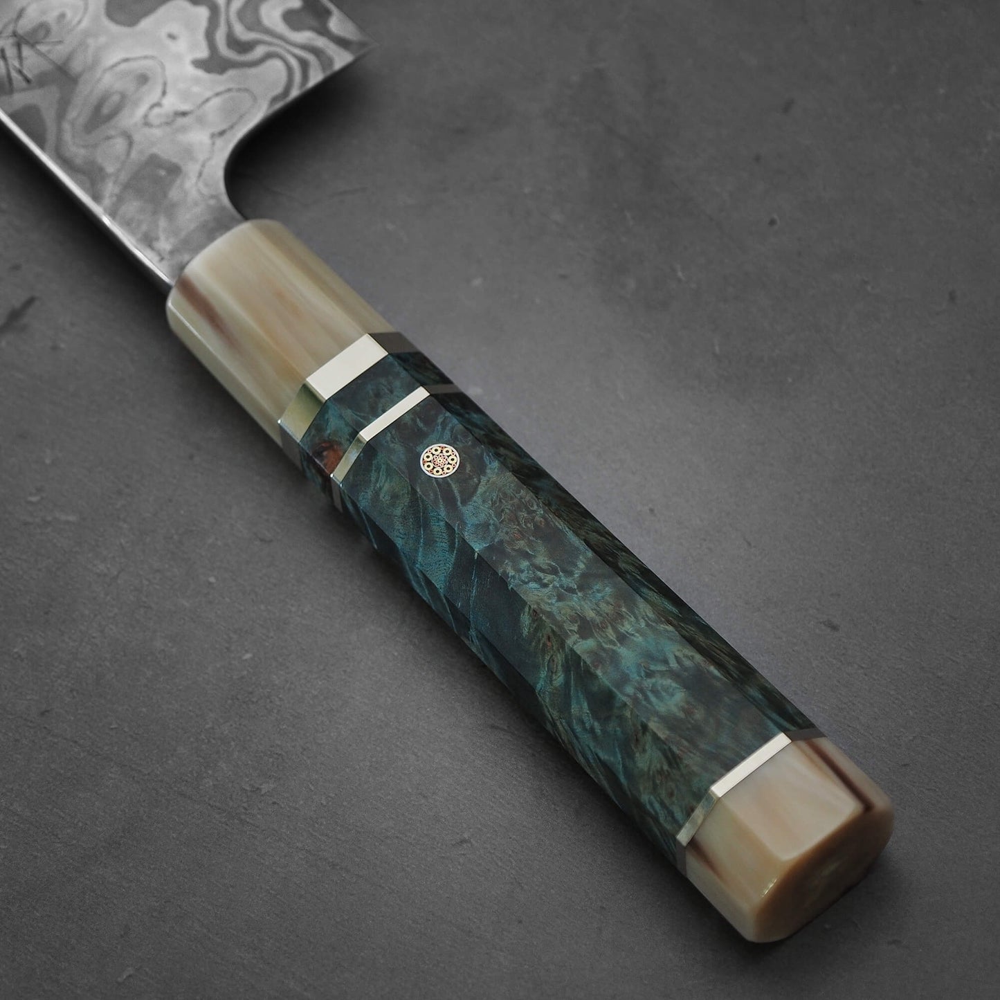 Close-up of the Nigara Hamono Nigara aogami#2 + shirogami#2 coreless honyaki gyuto 300mm knife, showcasing its Japanese sword-inspired patterned blade and a multicolored handle with metallic accents and a decorative round pin.