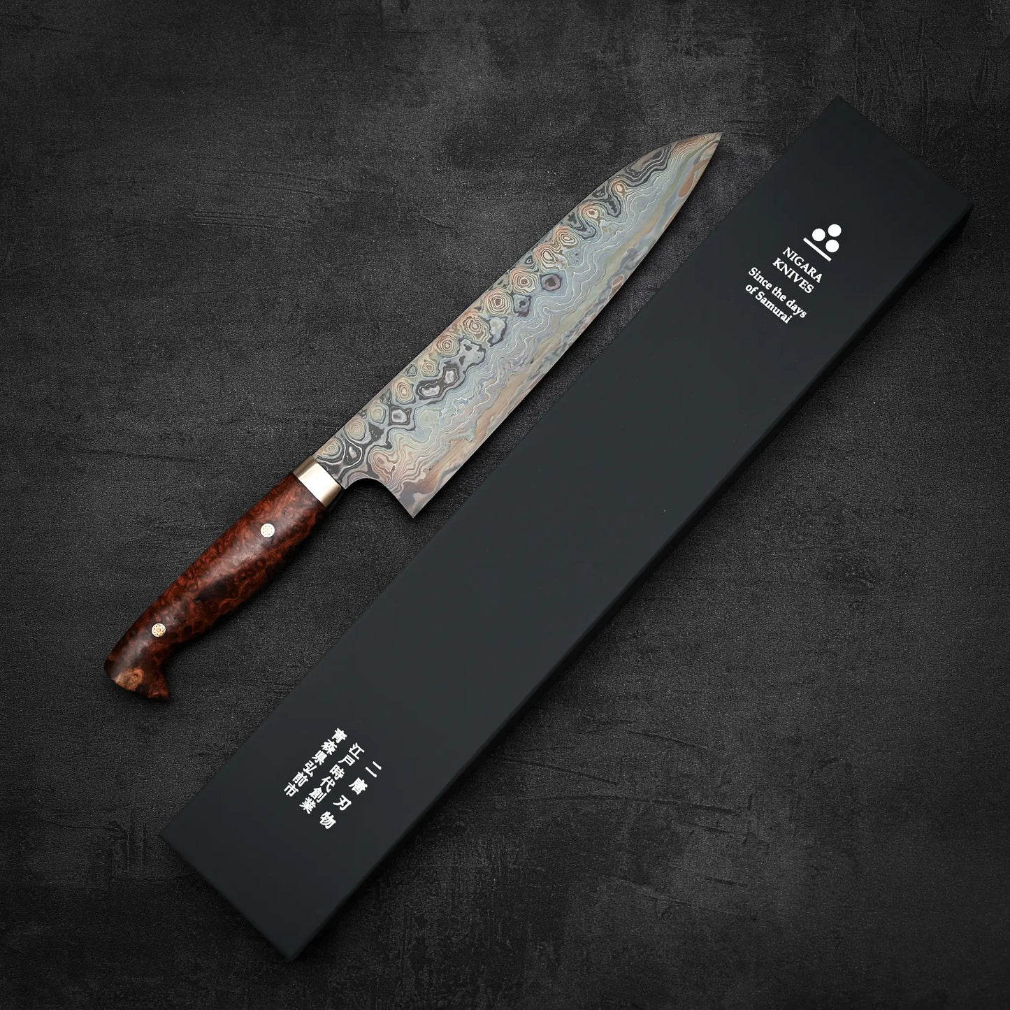A Nigara Hamono Nigara aogami#2 + shirogami#2 coreless damascus gyuto 240mm with a wooden handle rests beside a black box with white Japanese and English text, all arranged on a dark textured surface.