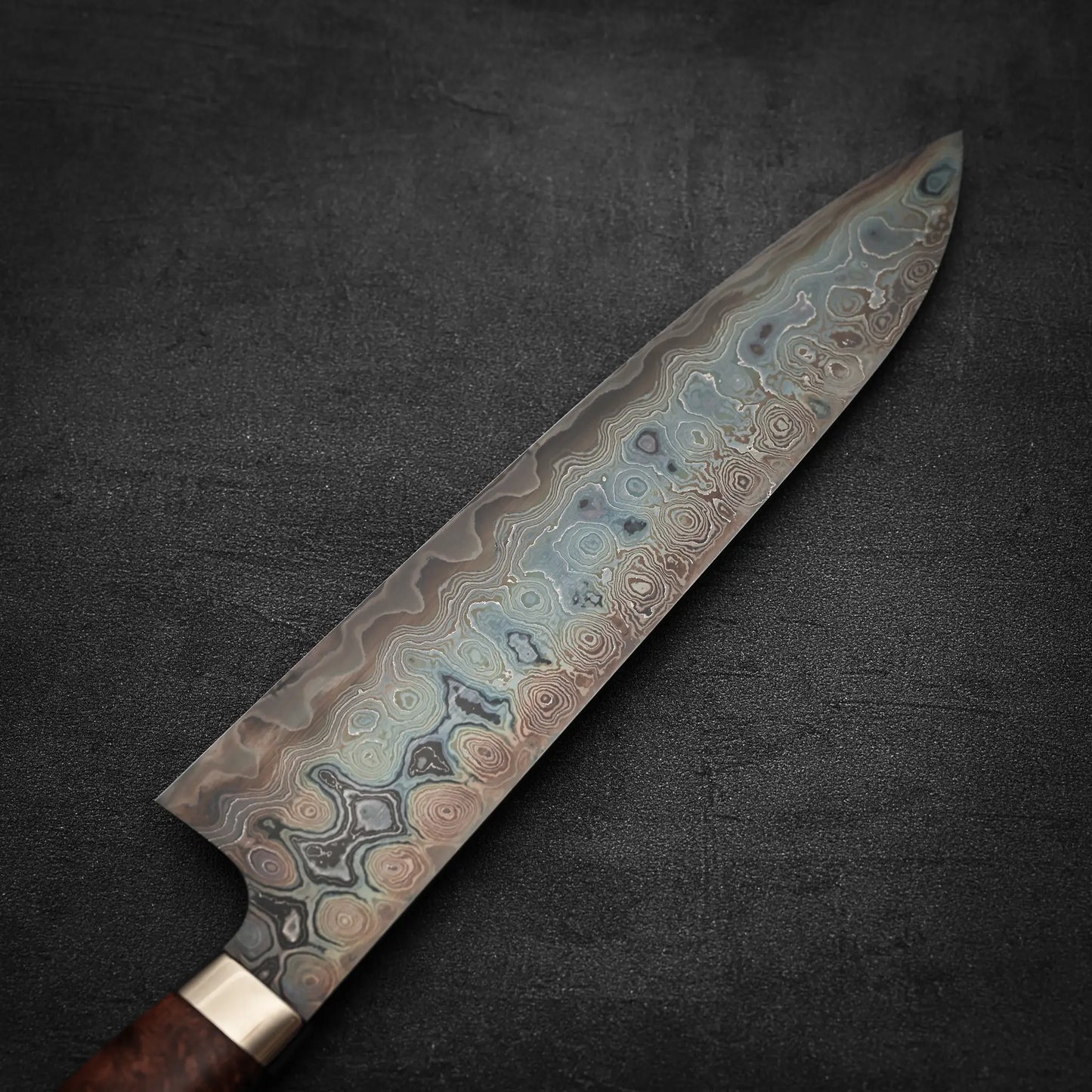 A close-up of the Nigara Hamono Nigara aogami#2 + shirogami#2 coreless damascus gyuto 240mm shows its wavy, layered Damascus steel blade and elegant handle on a dark textured surface.