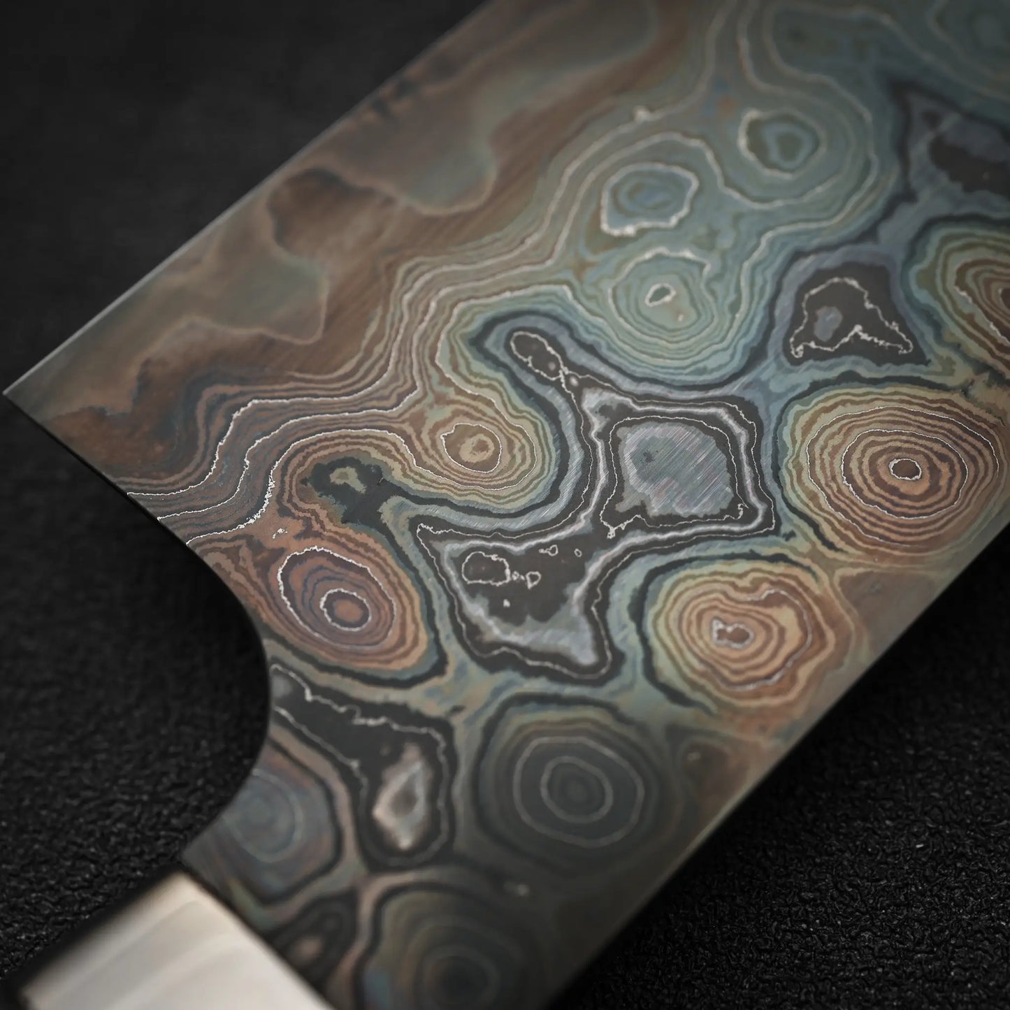 Close-up of the Nigara Hamono Nigara aogami#2 + shirogami#2 coreless damascus gyuto 240mm blade, highlighting its intricate, wavy layers in brown, gray, blue, and green against a textured black background.