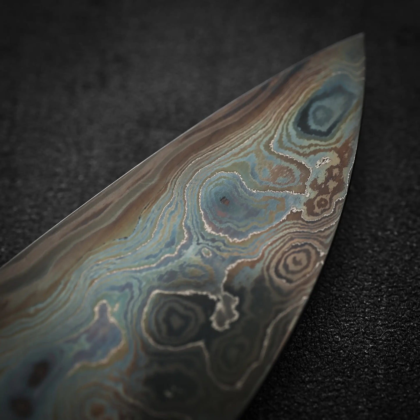 Close-up of the Nigara Hamono Nigara aogami#2 + shirogami#2 coreless damascus gyuto 240mm blade, showcasing its intricate, multicolored Damascus steel patterns with swirling blue, brown, and gray hues on a dark textured surface.