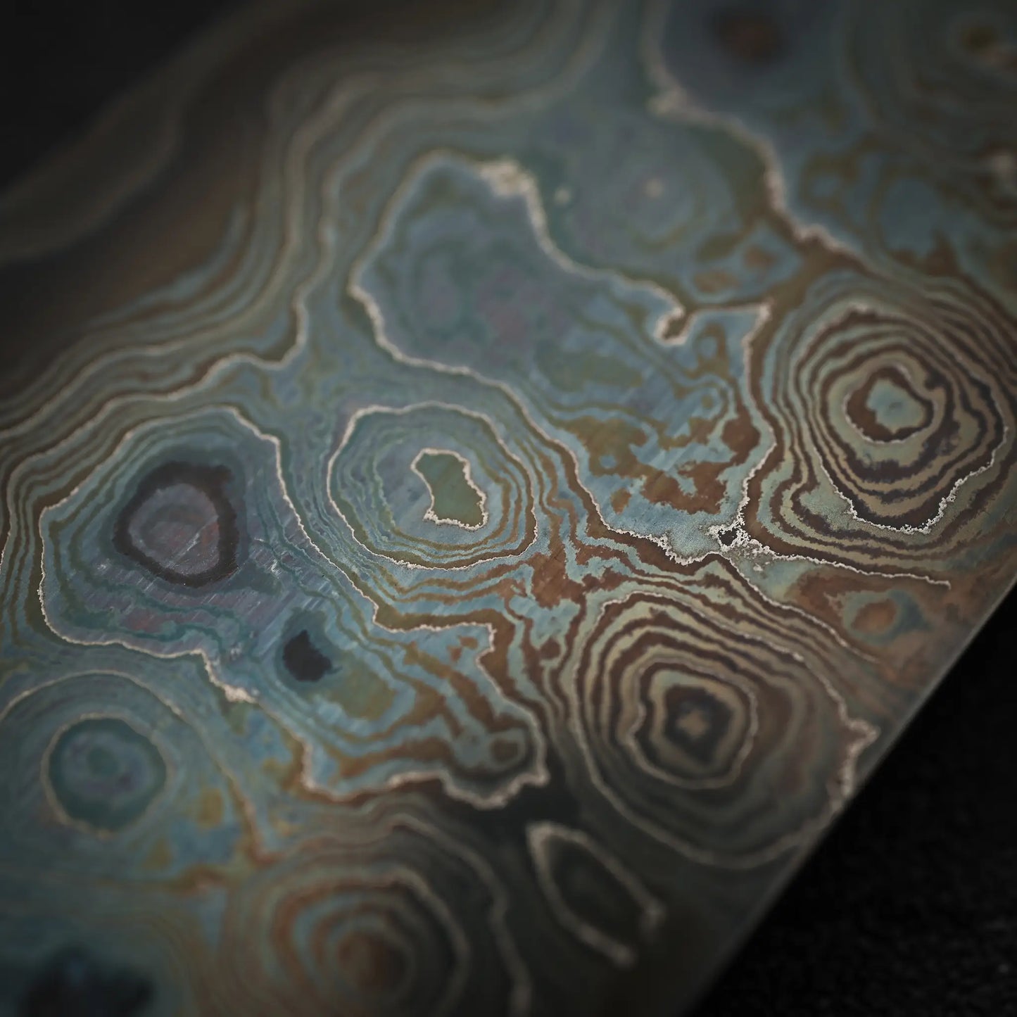 A close-up of the Nigara Hamono Nigara aogami#2 + shirogami#2 coreless damascus gyuto 240mm reveals wavy, layered blue, brown, and gray steel patterns reminiscent of intricate Damascus or gyuto blade designs.