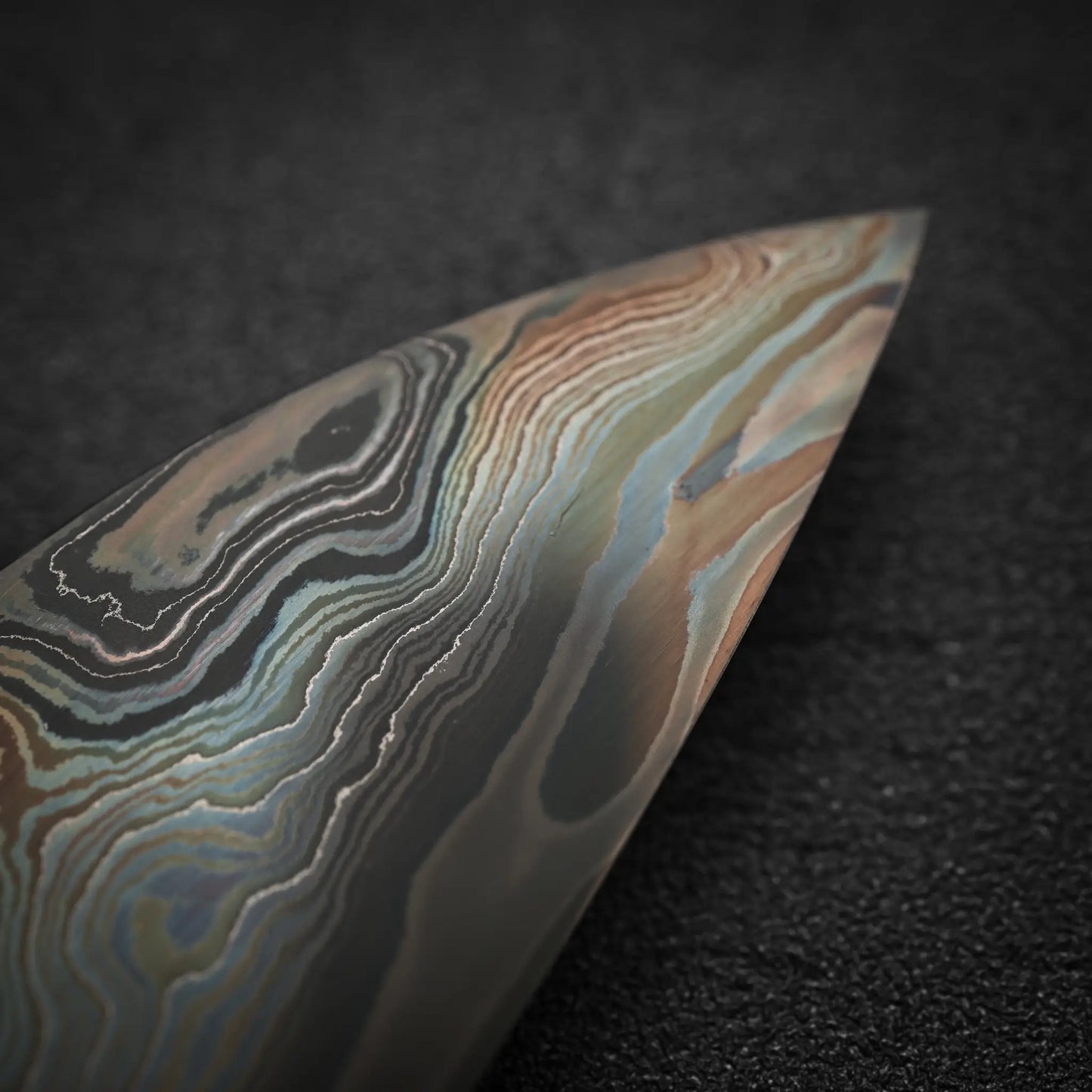 Close-up of the Nigara Hamono Nigara aogami#2 + shirogami#2 coreless damascus gyuto 240mm tip, its blade displaying intricate wavy damascus patterns against a dark, textured background.