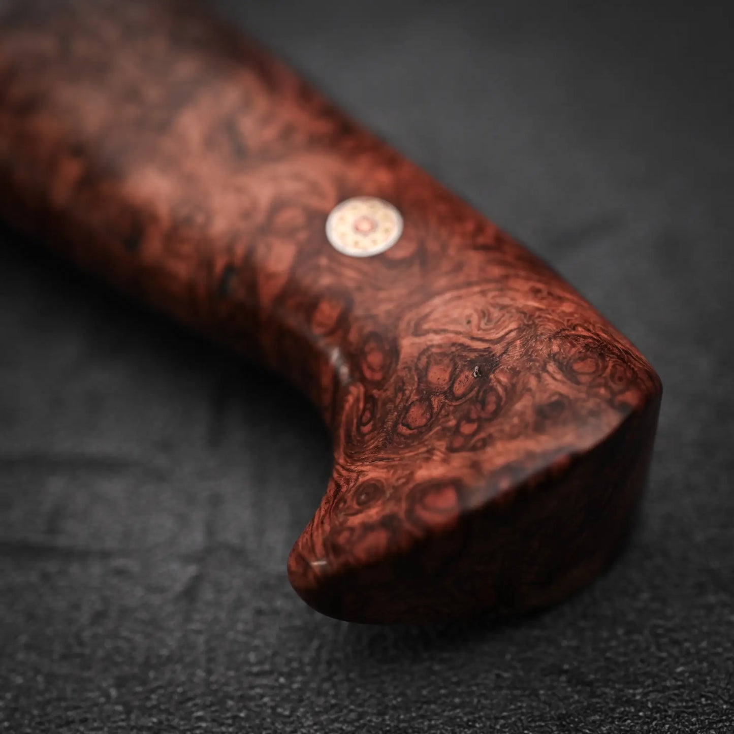 A close-up of the Nigara Hamono Nigara aogami#2 + shirogami#2 coreless damascus gyuto 240mm’s wooden handle, with a reddish-brown swirling grain and metal pin, set against a dark, textured background.