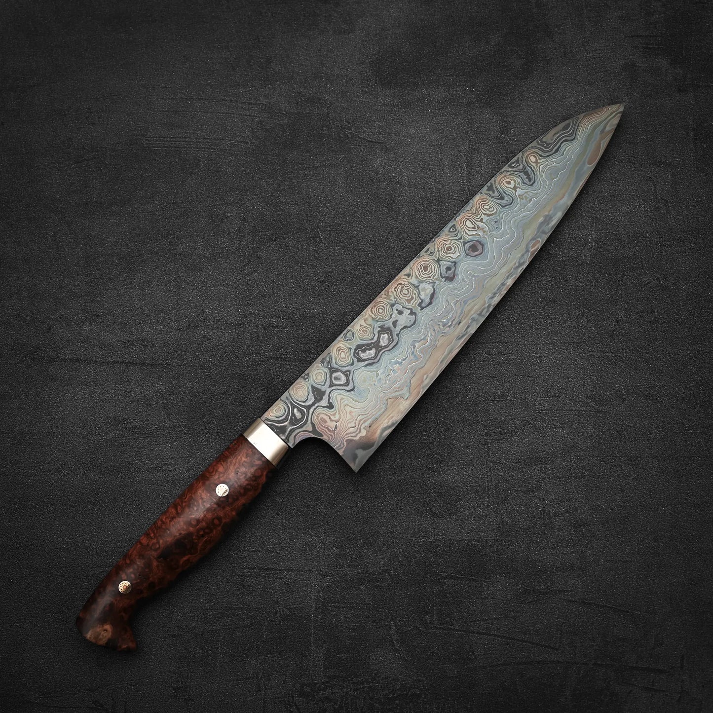 A Nigara Hamono aogami#2 + shirogami#2 coreless damascus gyuto 240mm chefs knife with a polished wood handle lies on a dark, textured surface.
