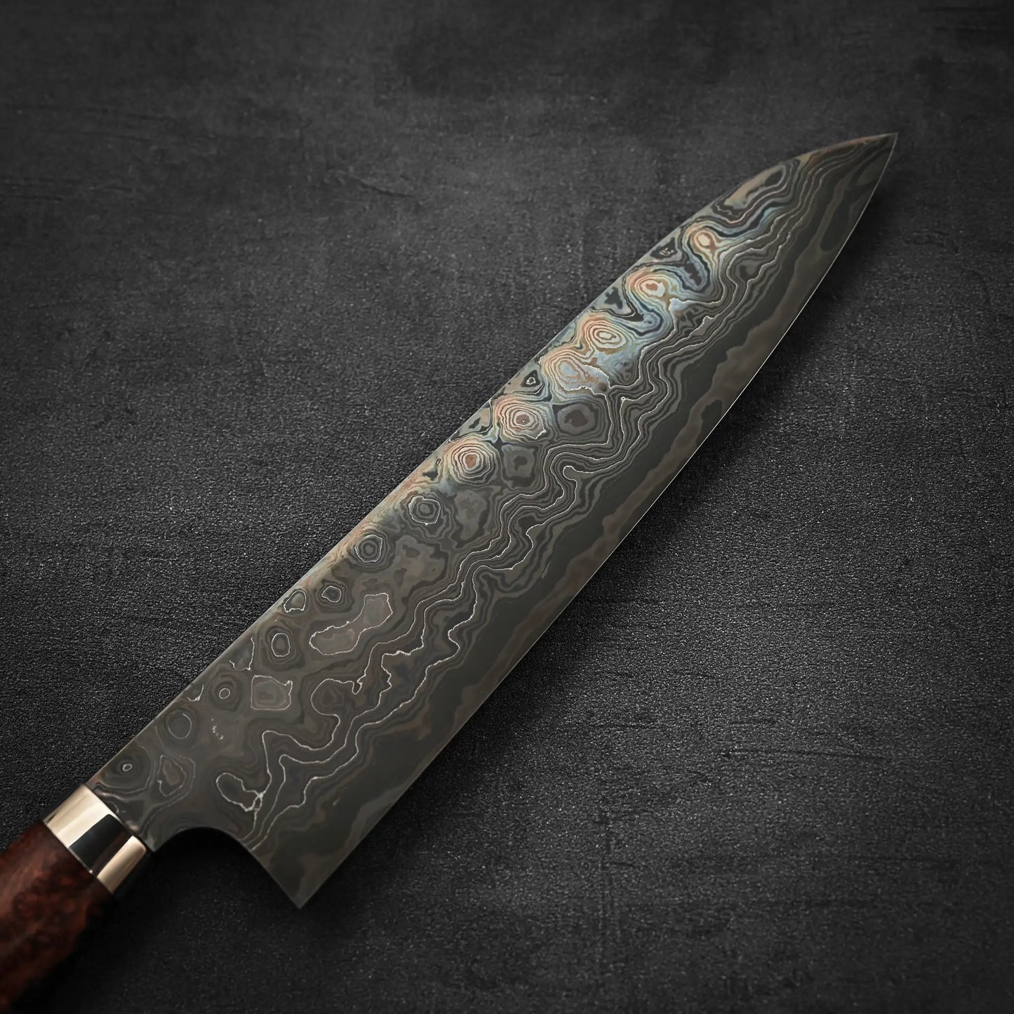 A close-up of the Nigara Hamono Nigara aogami#2 + shirogami#2 coreless damascus gyuto 240mm chefs knife with a dark damascus steel blade and wooden handle, resting on a textured dark gray surface.