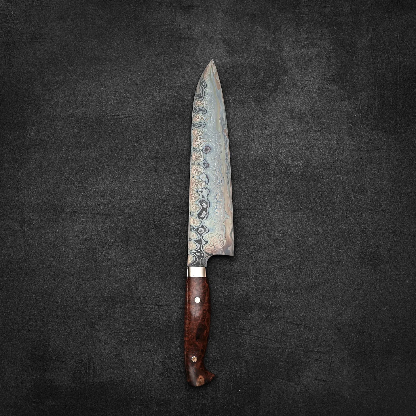 The Nigara Hamono Nigara aogami#2 + shirogami#2 coreless damascus gyuto 240mm, featuring a wavy damascus blade and polished wooden handle, rests on a dark, textured surface.