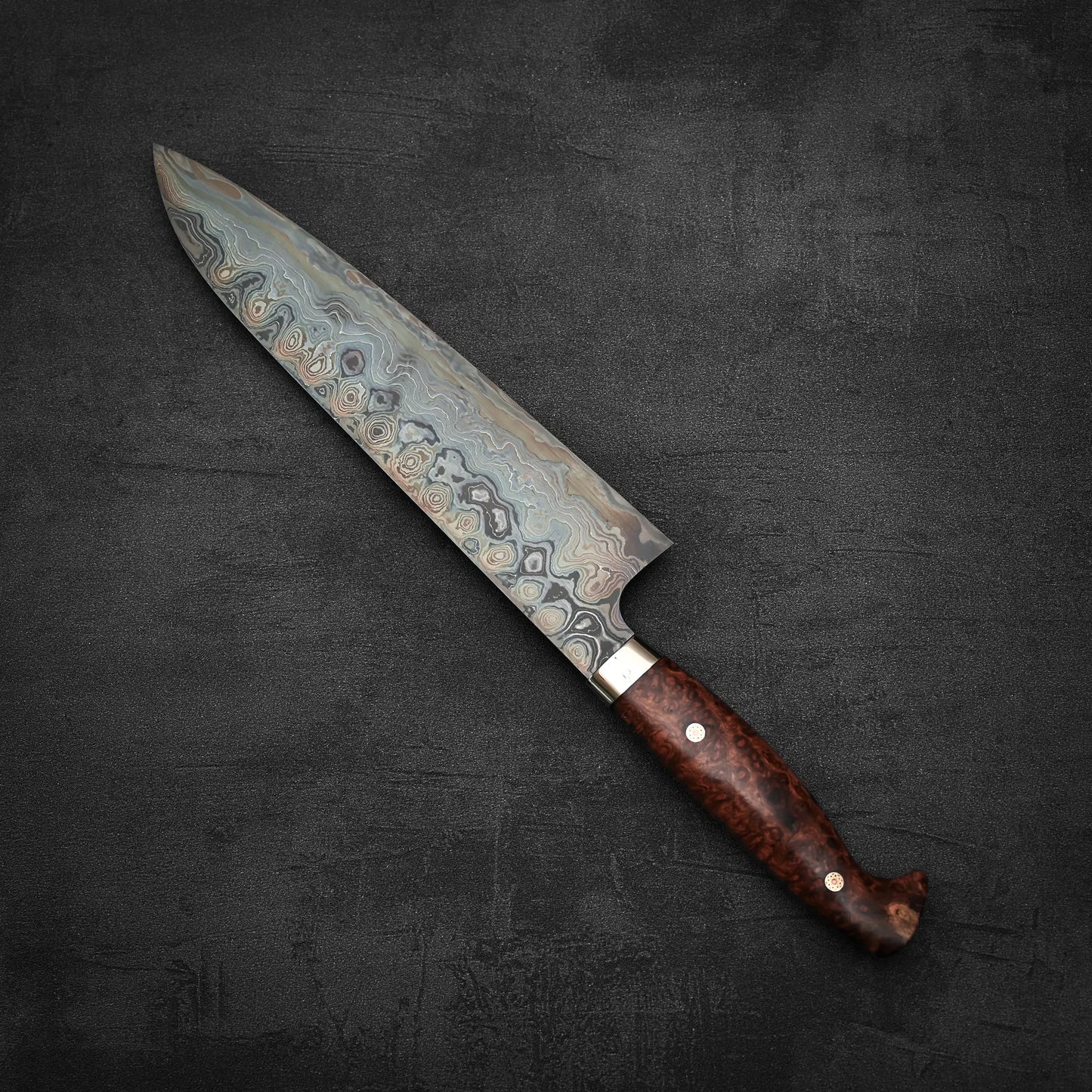 The Nigara Hamono Nigara aogami#2 + shirogami#2 coreless damascus gyuto 240mm, featuring a patterned Damascus steel blade and polished wooden handle, rests on a dark, textured surface.