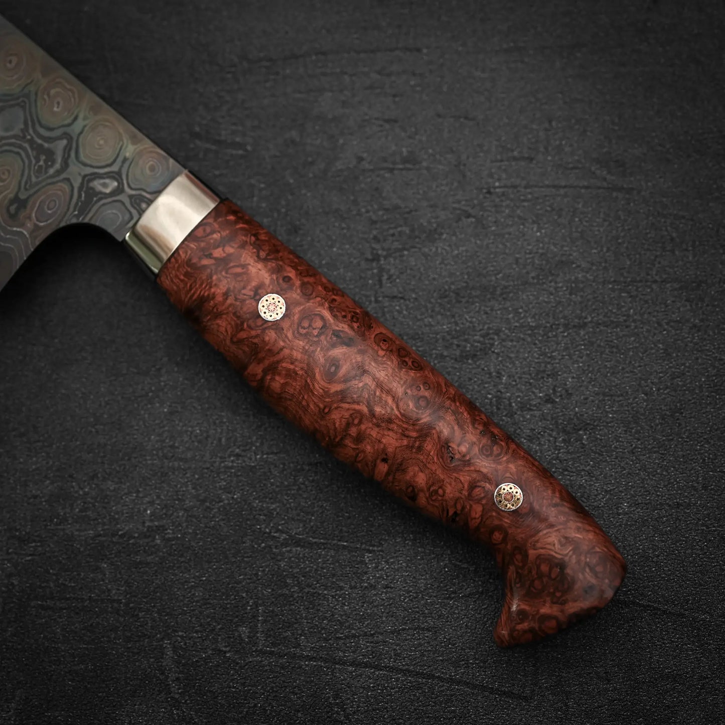 A close-up of the Nigara Hamono Nigara aogami#2 + shirogami#2 coreless damascus gyuto 240mm shows its polished, richly grained wooden handle with two metal pins and the base of its patterned blade against a dark textured background.