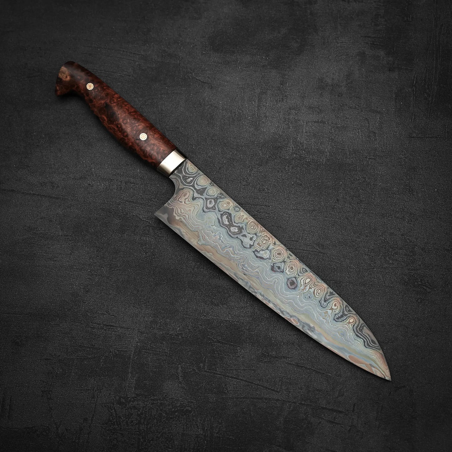 The Nigara Hamono Nigara aogami#2 + shirogami#2 coreless damascus gyuto 240mm, featuring a patterned Damascus steel blade and polished dark wooden handle, lies on a dark textured surface.
