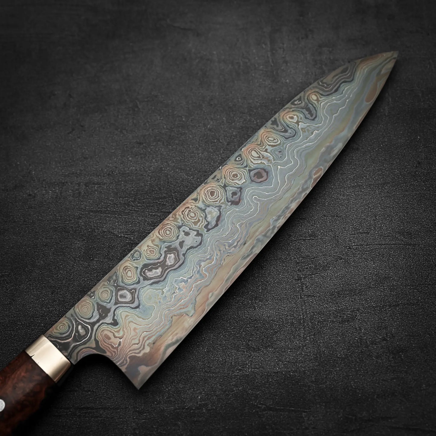 A close-up of the Nigara Hamono Nigara aogami#2 + shirogami#2 coreless damascus gyuto 240mm chef’s knife, featuring intricate wavy damascus patterns and a wooden handle, shown on a dark textured surface.