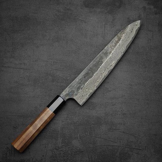 The Nigara Hamono Nigara Kurozome Damascus Ginsan Gyuto 240mm chef’s knife, featuring a wooden handle and patterned Damascus blade, rests on a dark textured surface, its wavy designs reflecting traditional Japanese artistry.