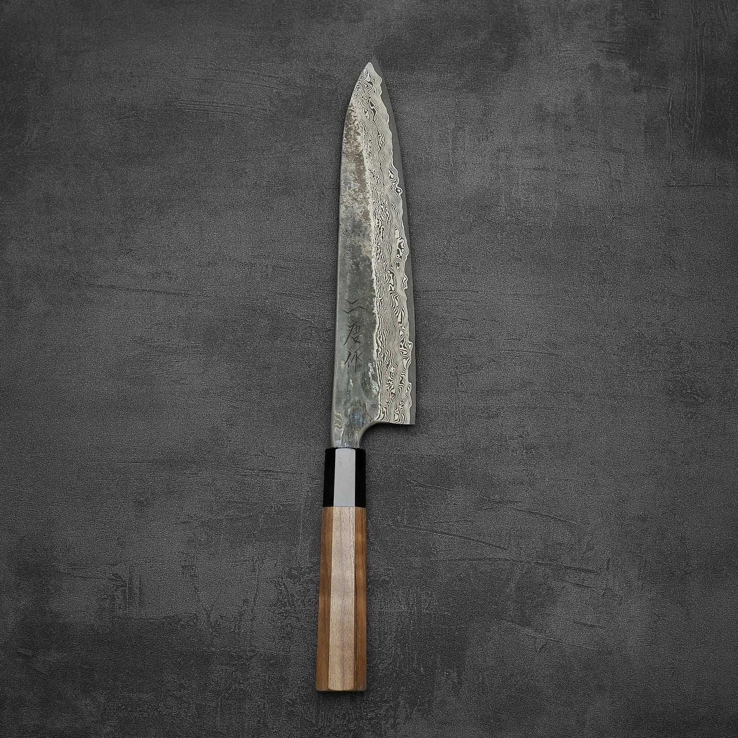 The Nigara Hamono Nigara Kurozome damascus ginsan gyuto 240mm, featuring a Damascus blade and wooden handle, rests on a dark, textured surface.
