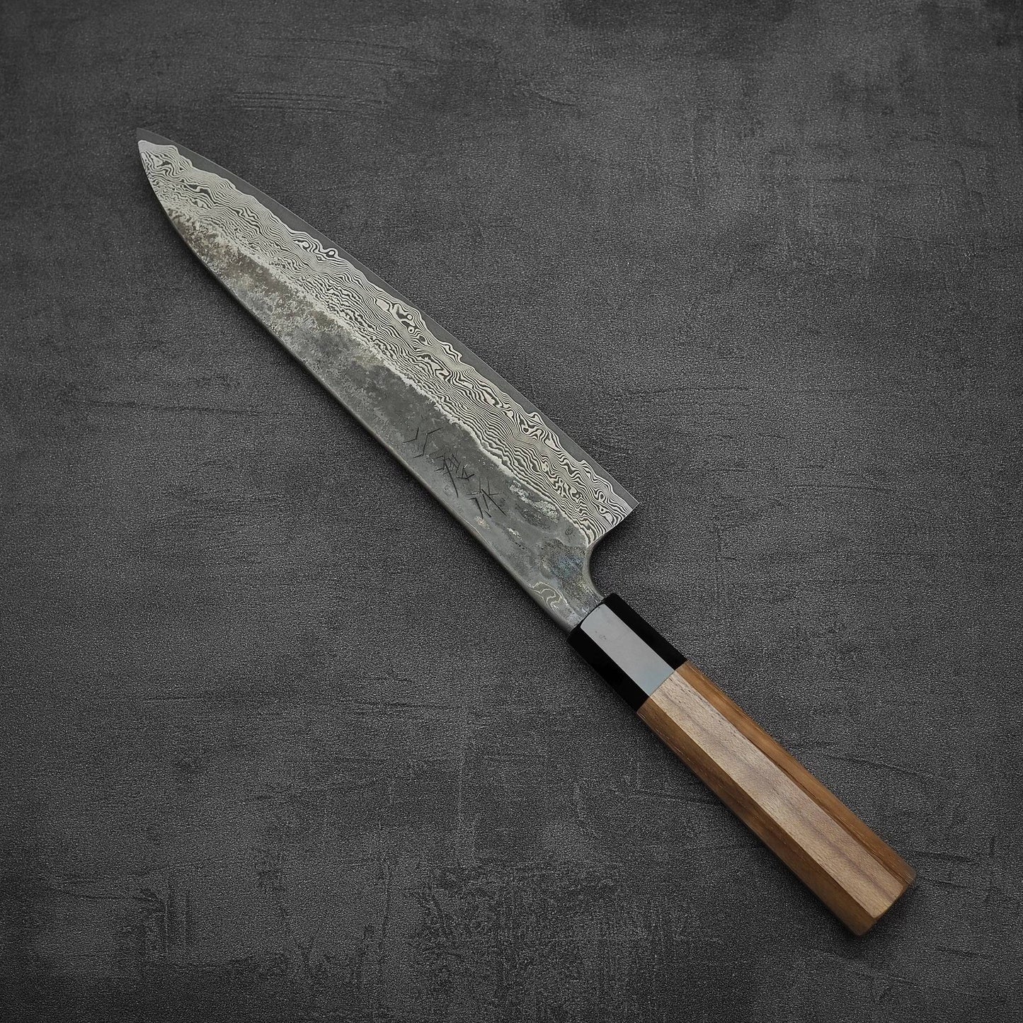 The Nigara Kurozome damascus ginsan gyuto 240mm from Nigara Hamono features a patterned Damascus blade and a wooden handle, set on a dark textured surface—ideal for those who appreciate elegant, precise traditional Japanese chef knives.