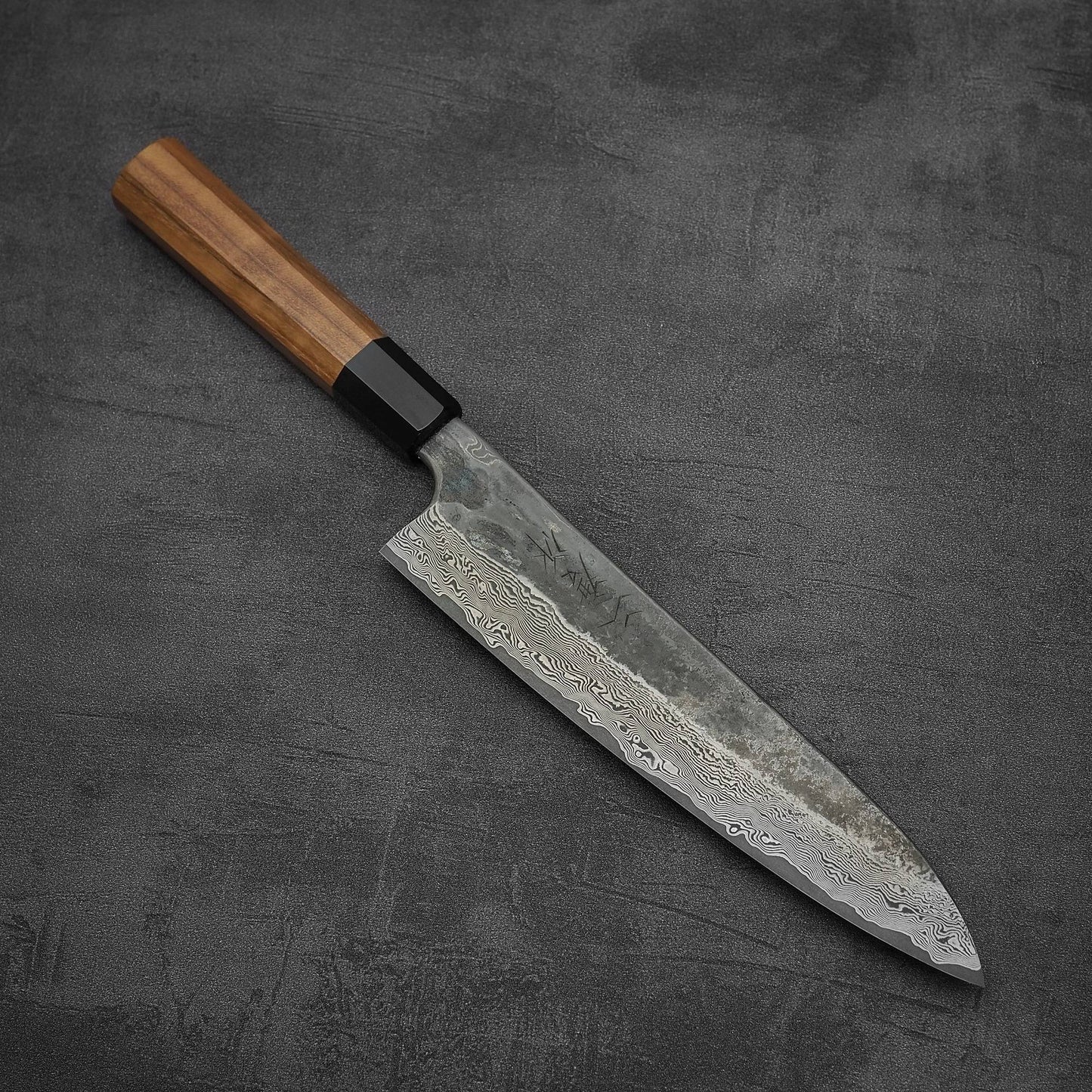 The Nigara Hamono Nigara Kurozome damascus ginsan gyuto 240mm, with its Damascus blade and wooden handle, lies on a dark textured surface, highlighting the craftsmanship of Japanese knives.