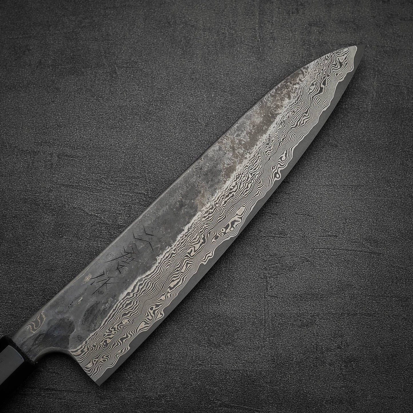 A close-up of the Nigara Hamono Nigara Kurozome damascus ginsan gyuto 240mm, showcasing its dark handle and patterned Damascus blade, inspired by Japanese knives, resting on a textured dark surface.