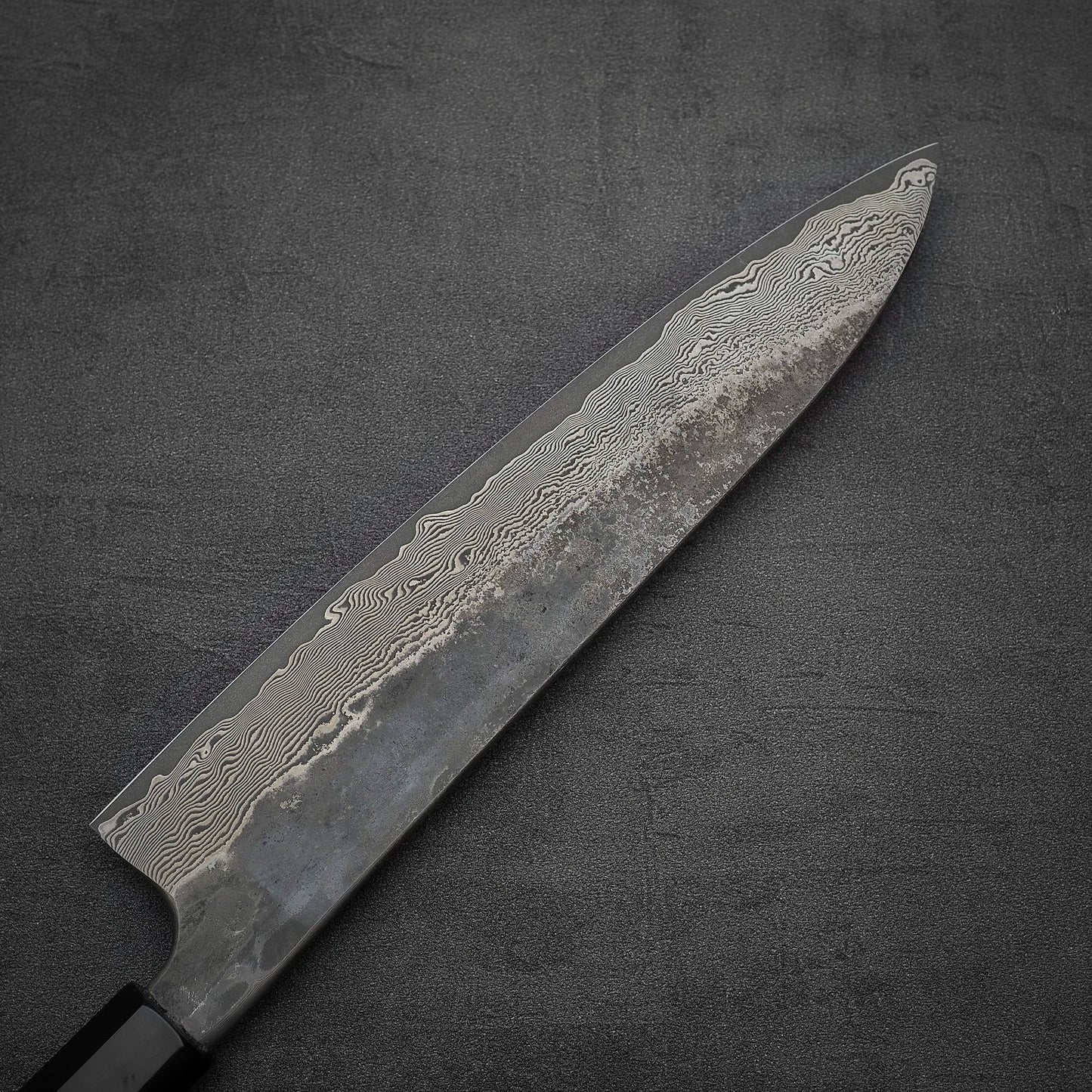 A close-up of the Nigara Hamono Nigara Kurozome damascus ginsan gyuto 240mm kitchen knife, showcasing its distinctive Damascus wavy patterns on a dark, textured surface.