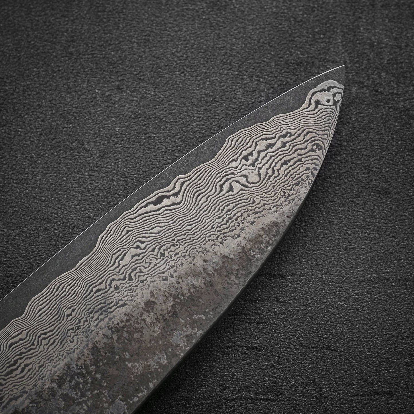 Close-up of the Nigara Hamono Nigara Kurozome damascus ginsan gyuto 240mm tip, highlighting its intricate wavy Damascus patterns and textured metal, reminiscent of Japanese knives, set against a dark background.