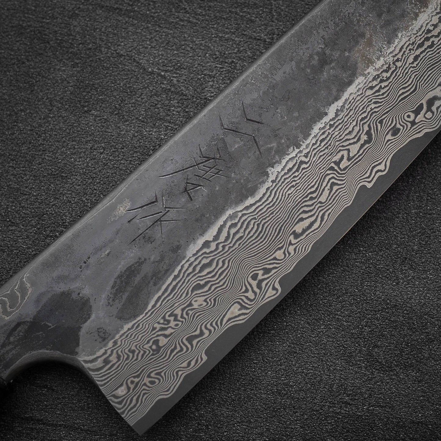 Close-up of the Nigara Hamono Nigara Kurozome damascus ginsan gyuto 240mm knife, showcasing Ginsan steel with etched wavy damascus lines and Japanese-style characters on a dark textured background.