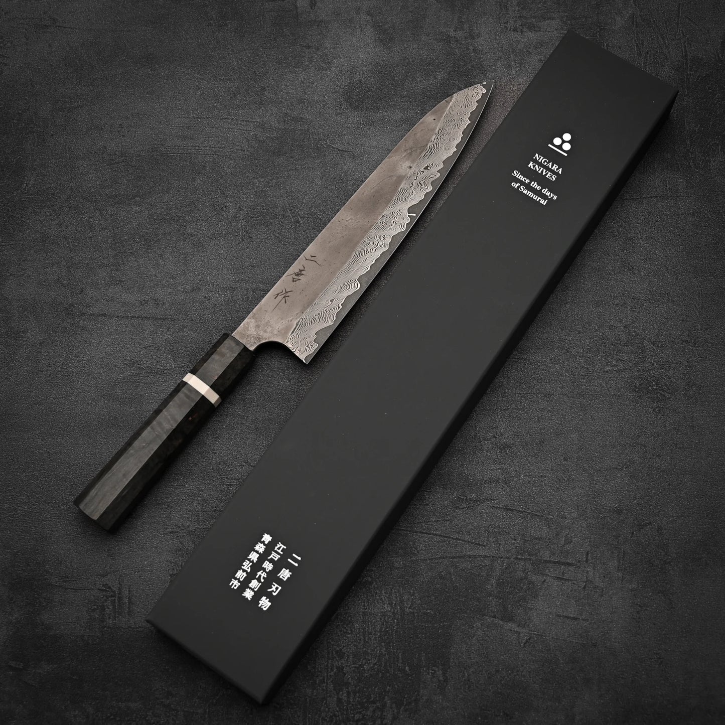 The Nigara Hamono Nigara Kurozome damascus ginsan gyuto 240mm chefs knife with a dark wooden handle and patterned blade sits beside a black box with white English and Japanese text on a dark textured surface.