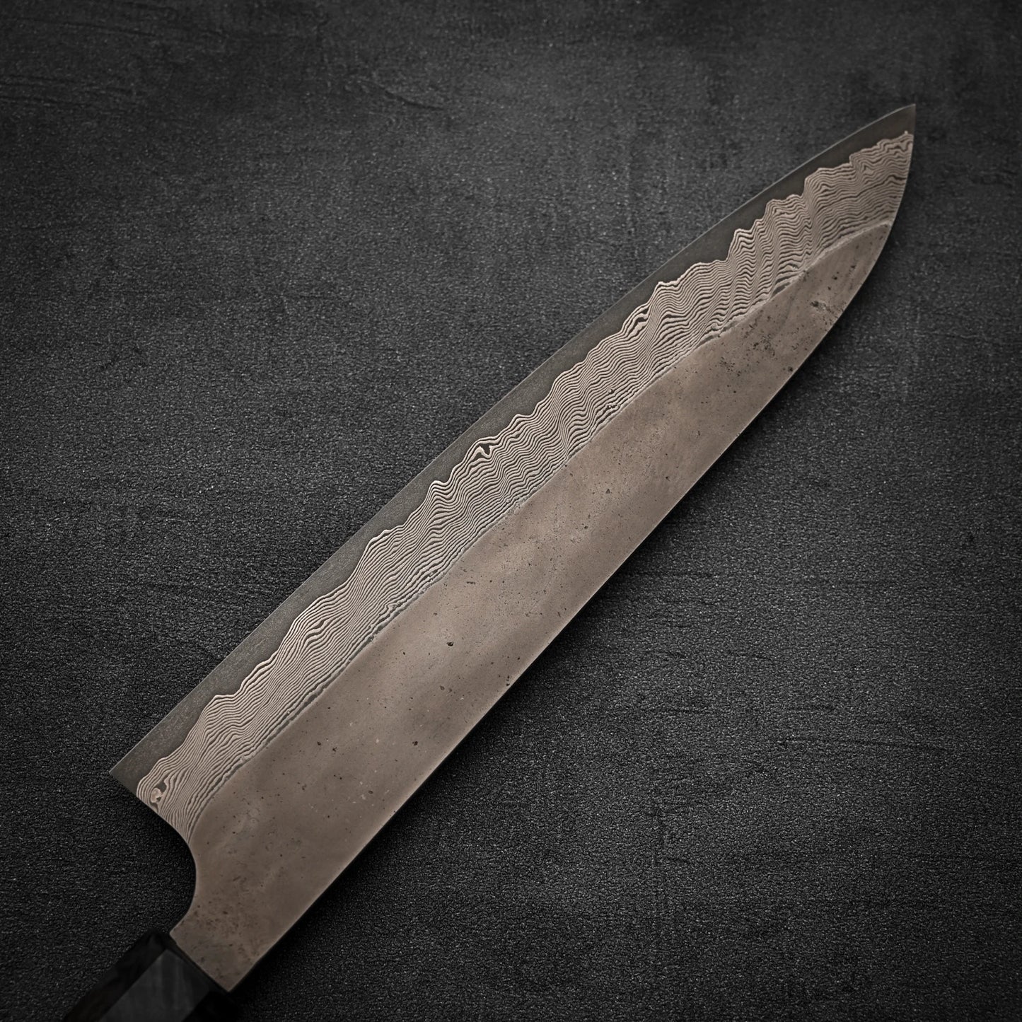 A close-up of the Nigara Hamono Nigara Kurozome damascus ginsan gyuto 240mm knife, showcasing its patterned wavy Damascus blade against a dark textured background.