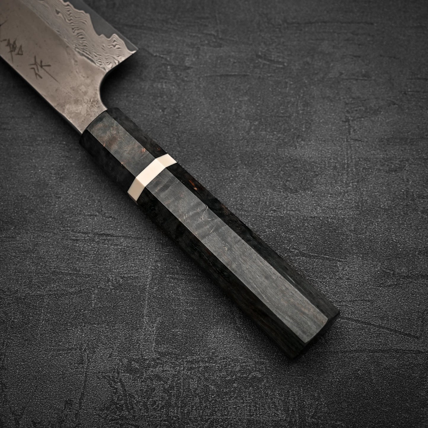 Close-up of the Nigara Hamono Nigara Kurozome damascus ginsan gyuto 240mm, showing its dark octagonal wooden handle with a light ring near the blade and part of its patterned damascus ginsan steel blade on a dark surface.