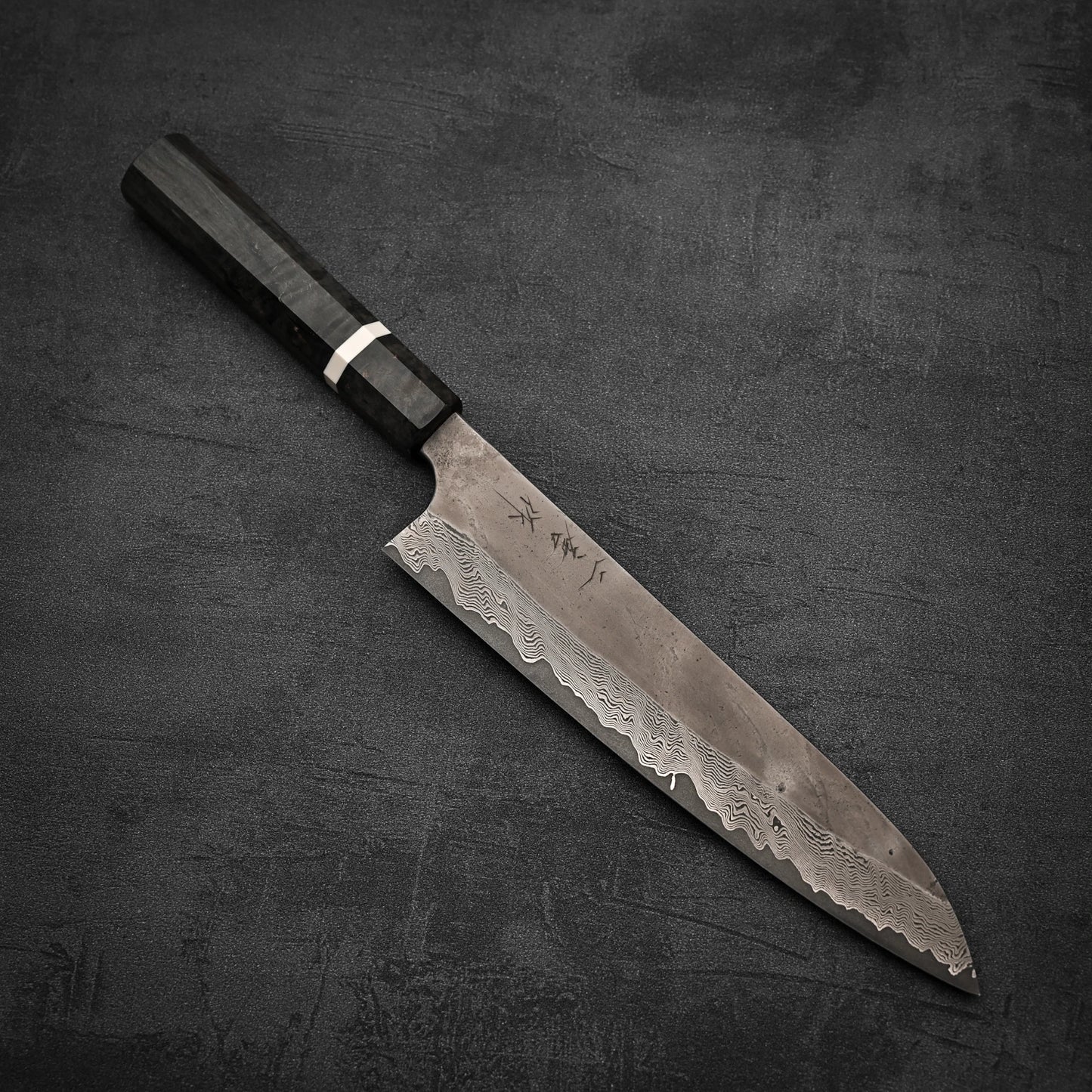 The Nigara Hamono Nigara Kurozome damascus ginsan gyuto 240mm, featuring a dark wood handle and shimmering damascus blade, rests elegantly on a textured dark gray surface.