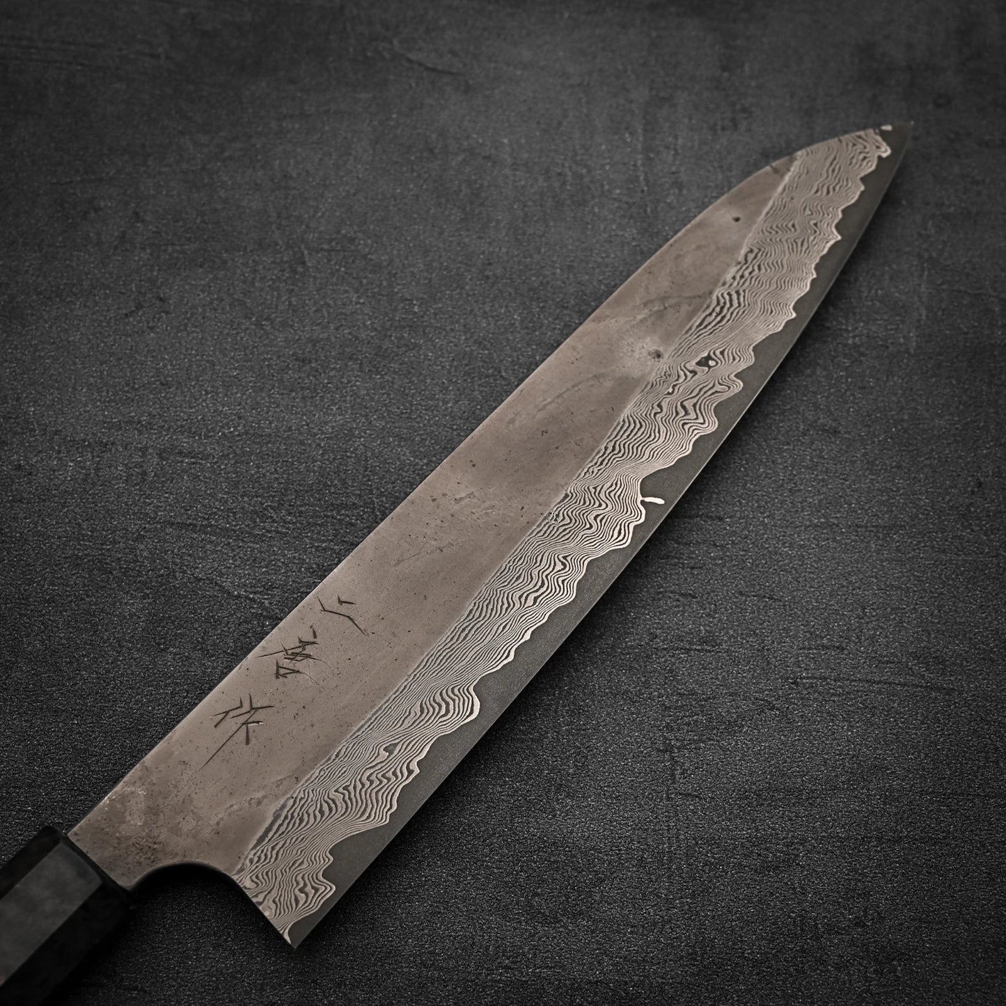 A close-up of the Nigara Hamono Nigara Kurozome Damascus Ginsan Gyuto 240mm, showing its intricate Damascus steel patterns and engraved Japanese characters on the blade, set against a dark textured background.