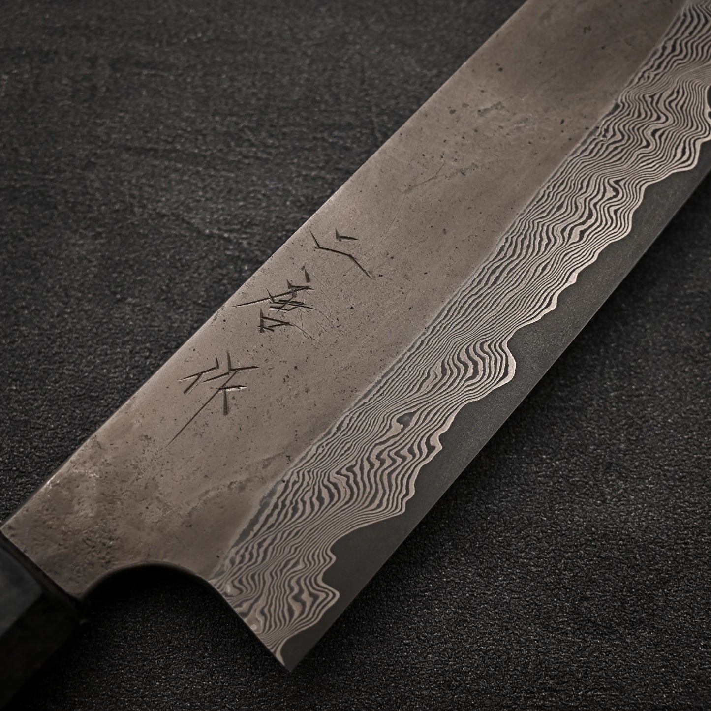 Close-up of the Nigara Hamono Nigara Kurozome damascus ginsan gyuto 240mm knife blade, featuring intricate wavy patterns and etched markings near the handle, set on a textured dark surface.
