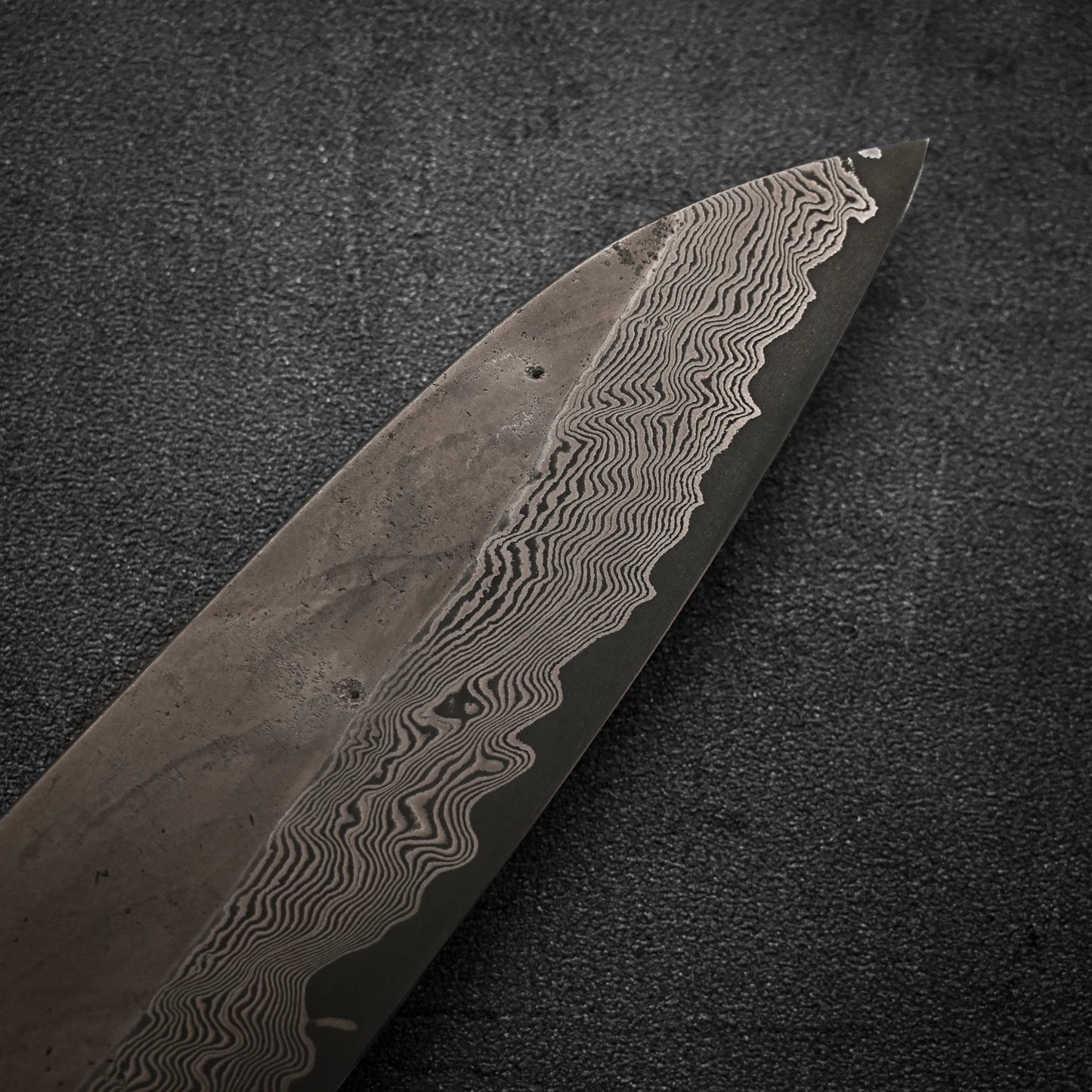 Close-up of the Nigara Hamono Nigara Kurozome damascus ginsan gyuto 240mm, showing its distinctive Damascus wavy patterns on the blade against a dark textured background, with intricate lines visible along the edge.