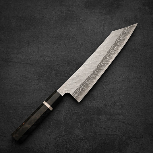 The Nigara Hamono Nigara tsuchime damascus VG10 kiritsuke gyuto 240mm—featuring a patterned VG10 steel blade and dark angular handle—rests on a textured dark surface.