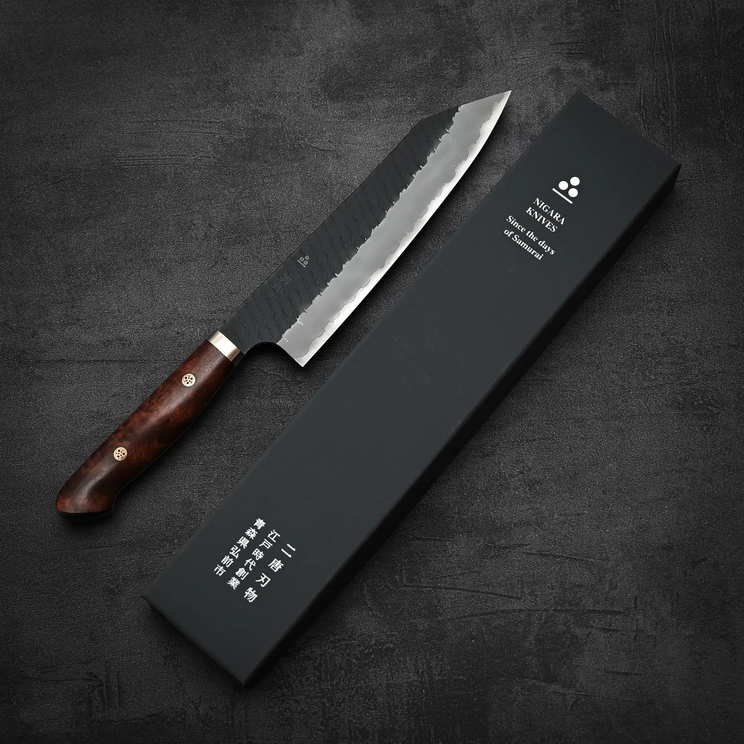 A Nigara Hamono Nigara tsuchime kurouchi SG2 kiritsuke gyuto 210mm chef’s knife with a dark wood handle and hammered blade sits beside a black knife box with Japanese and English text on a dark, textured surface.