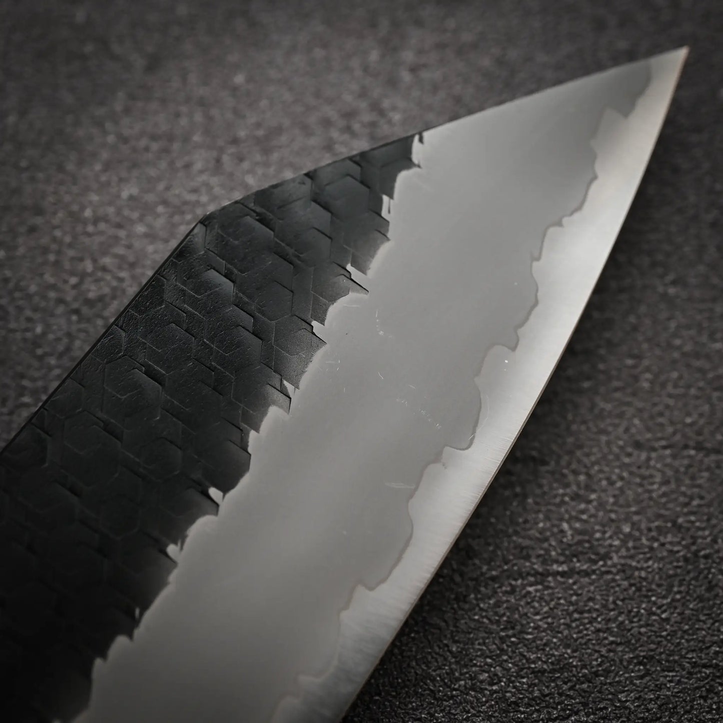 Close-up of the Nigara Hamono Nigara tsuchime kurouchi SG2 kiritsuke gyuto 210mm, highlighting its wavy blade pattern and textured dark spine, displayed on a rugged, dark surface.