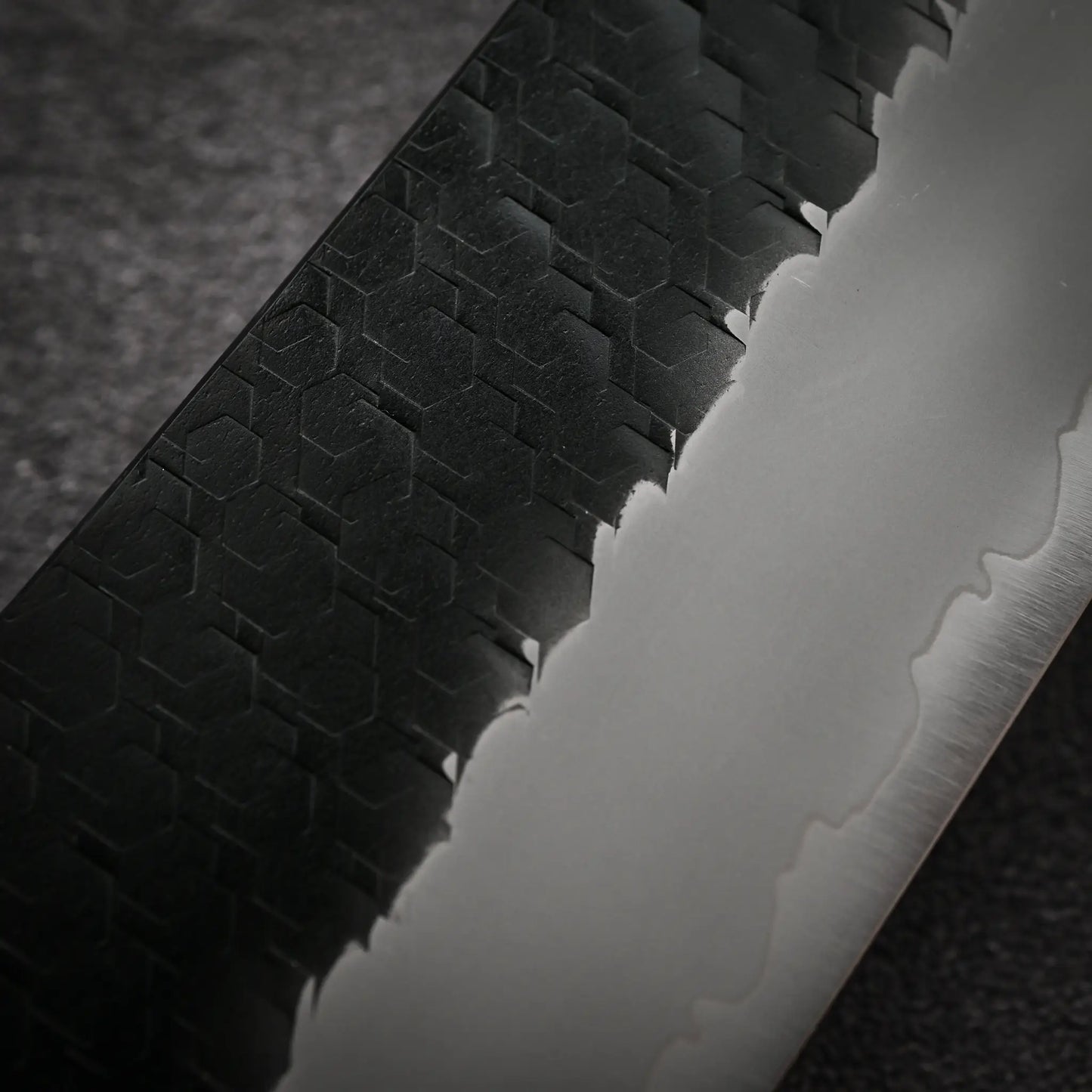 Close-up of the Nigara Hamono Nigara tsuchime kurouchi SG2 kiritsuke gyuto 210mm chef’s knife, showing its matte black blade with hexagonal patterns and silvery edge, detailed against a dark textured background.