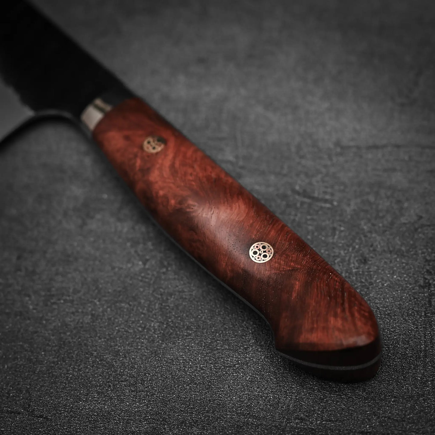 Close-up of the Nigara Hamono Nigara tsuchime kurouchi SG2 kiritsuke gyuto 210mm knifes polished dark wood handle with two metal rivets, resting on a textured black surface.