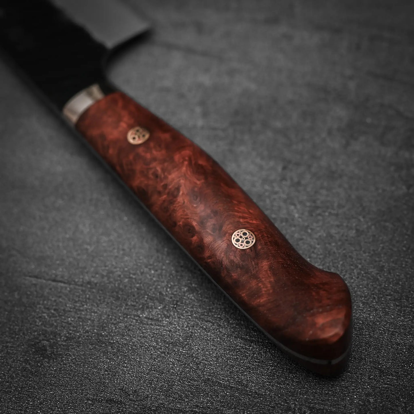 A close-up of the Nigara Hamono Nigara tsuchime kurouchi SG2 kiritsuke gyuto 210mm shows its polished reddish-brown wooden handle with two brass pins, resting on a dark textured surface; the blade appears partially at the top left.