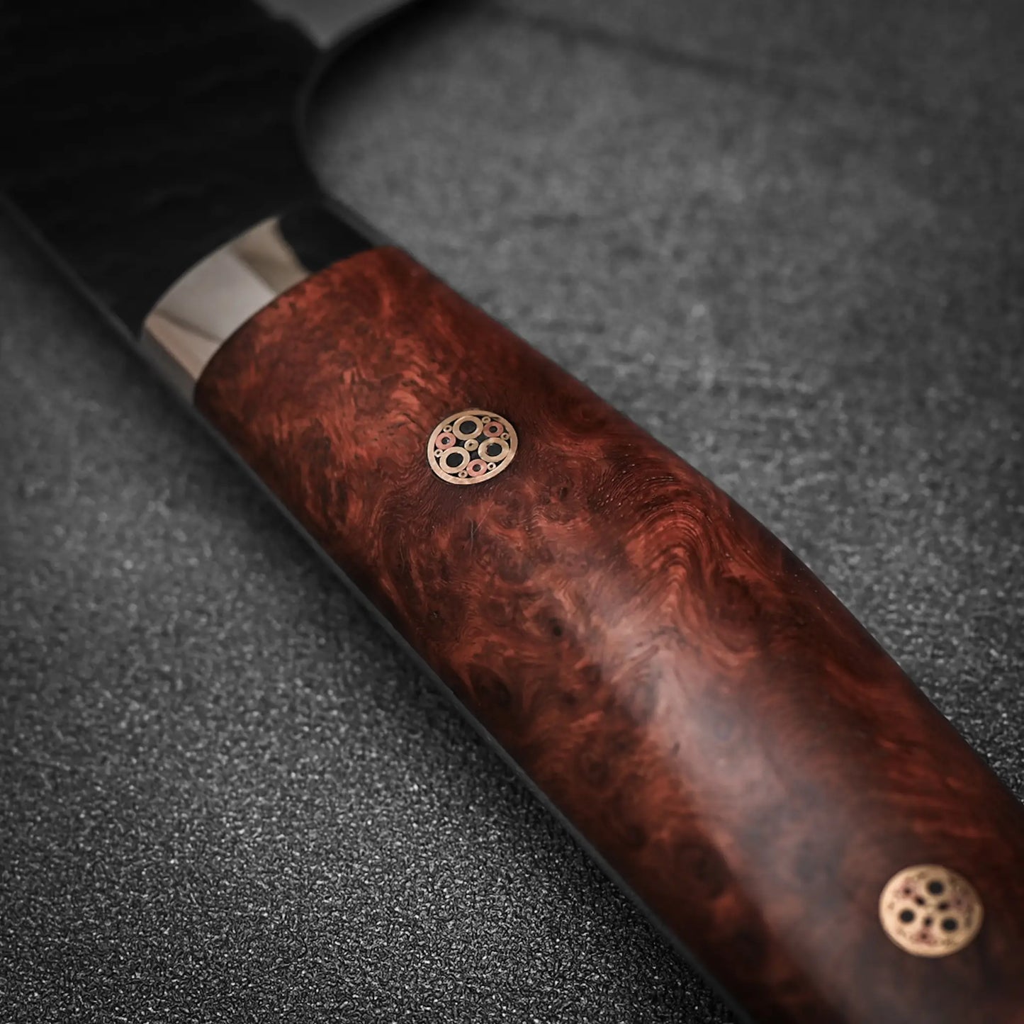 Close-up of the Nigara Hamono Nigara tsuchime kurouchi SG2 kiritsuke gyuto 210mm knife handle, crafted from polished dark wood with two metal rivets, shown resting on a textured dark surface.