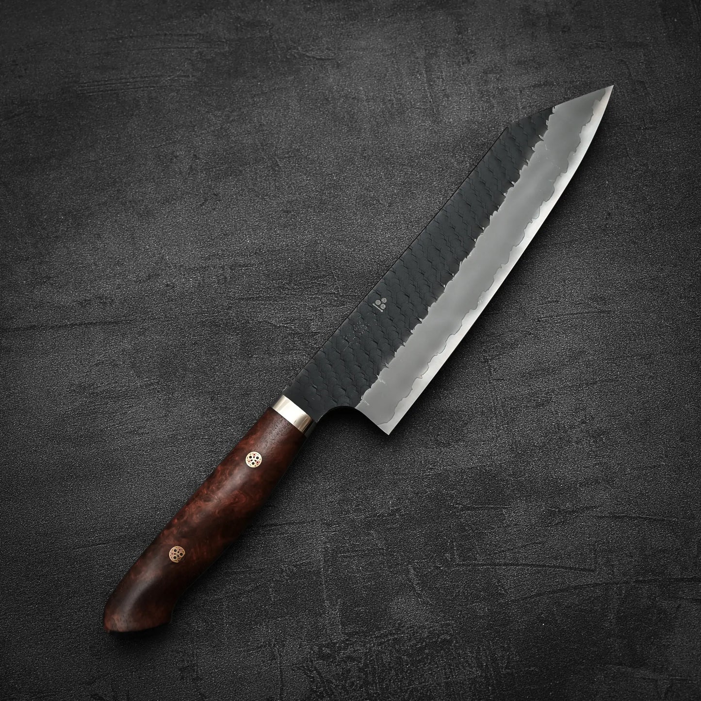 The Nigara Hamono Nigara tsuchime kurouchi SG2 kiritsuke gyuto 210mm, featuring a dark wooden handle and hammered blade, rests on a dark gray textured surface.